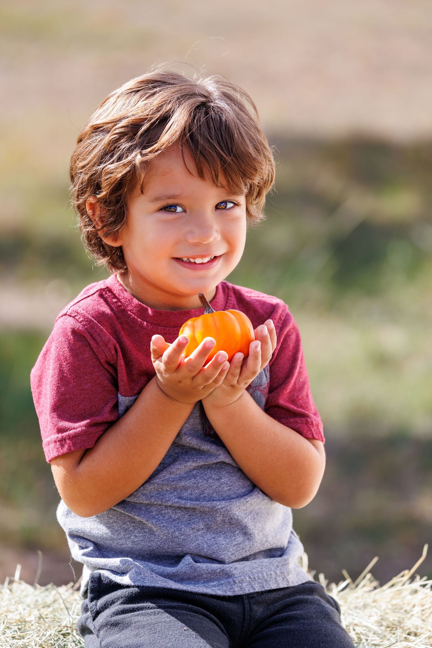 Young child holding an orange pumpkin while sitting on hay bales outdoors.
