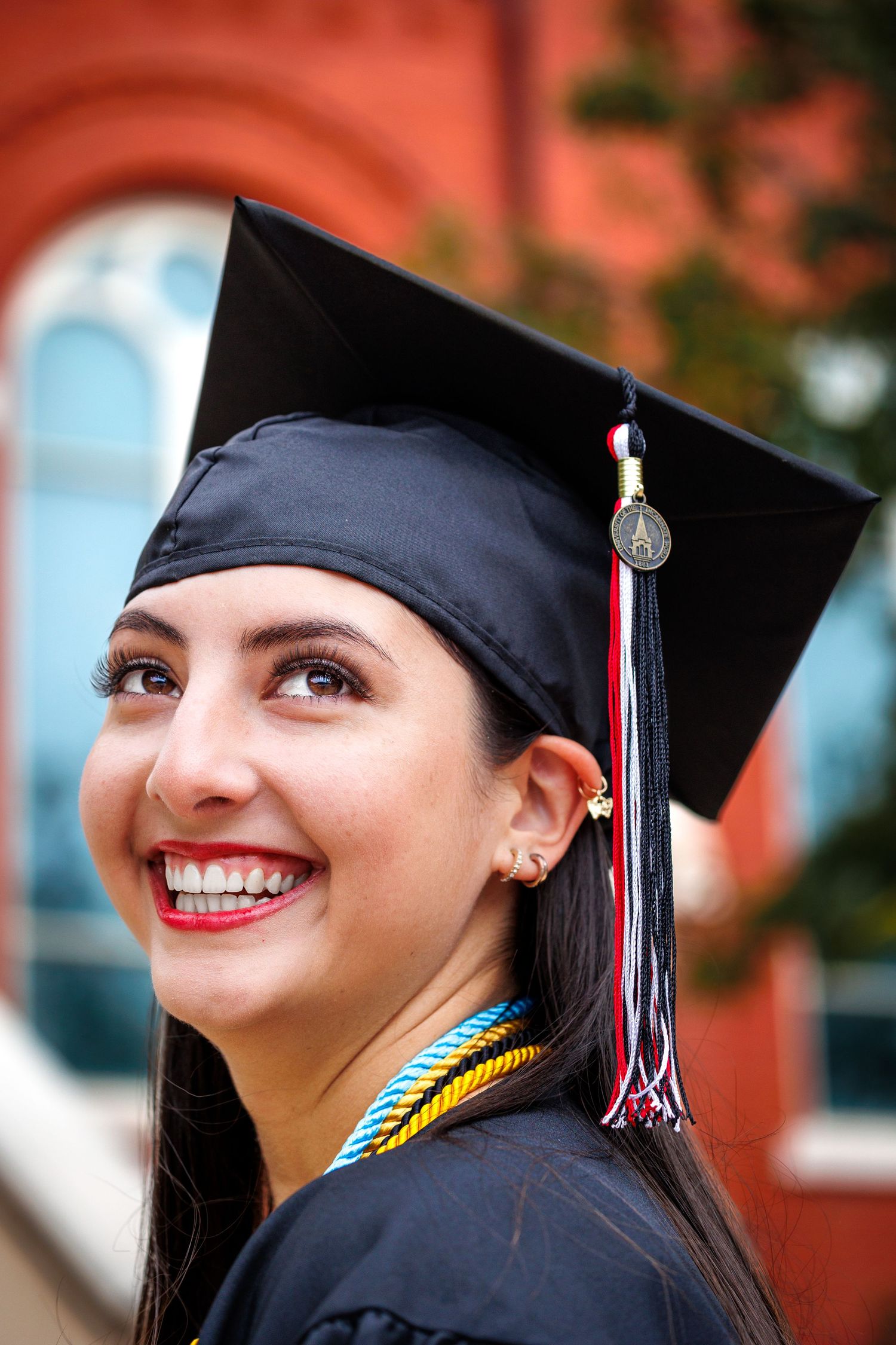 Graduate in cap and gown with tassel smiling against red brick building.
