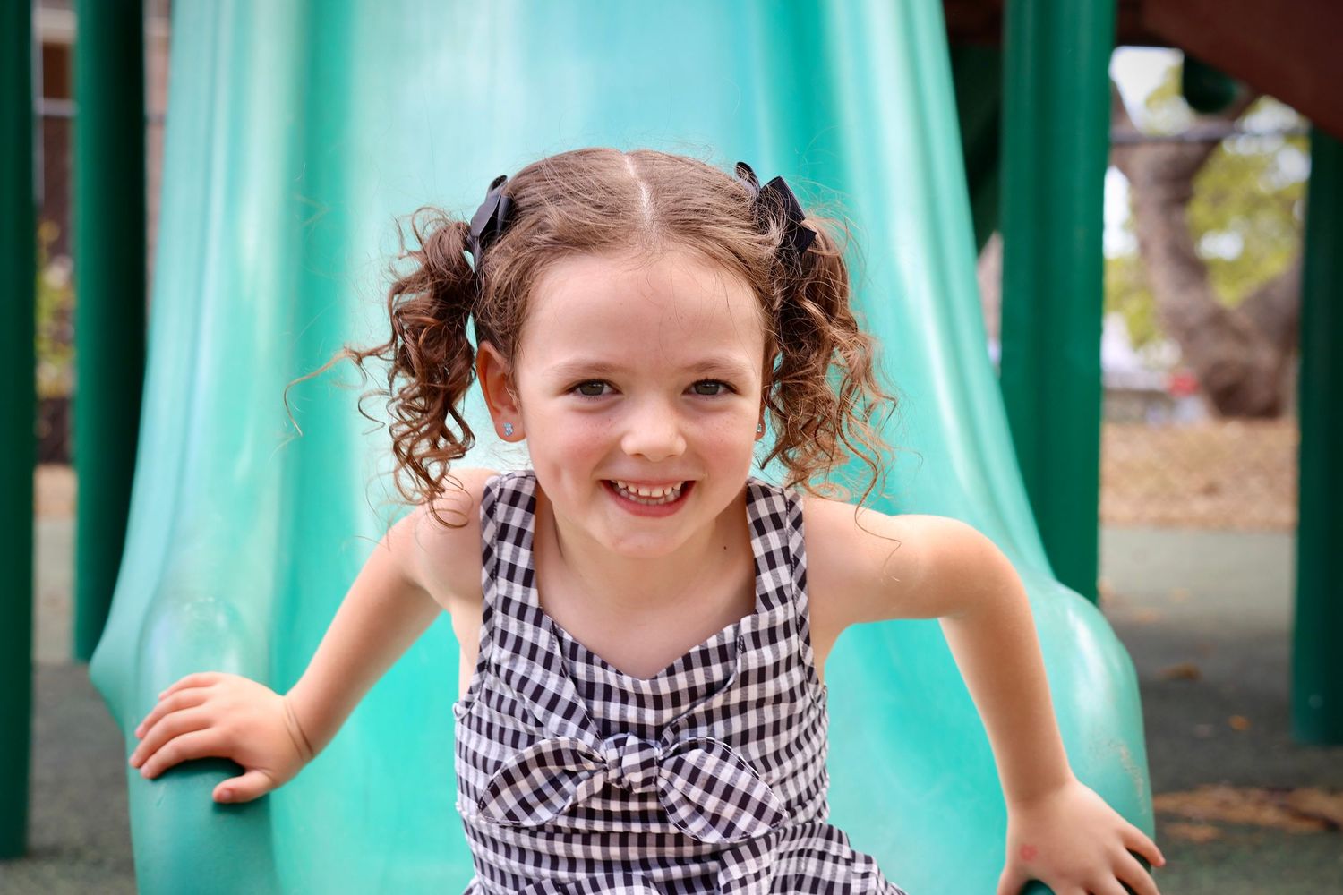 Young child in checkered dress playing on turquoise playground slide.