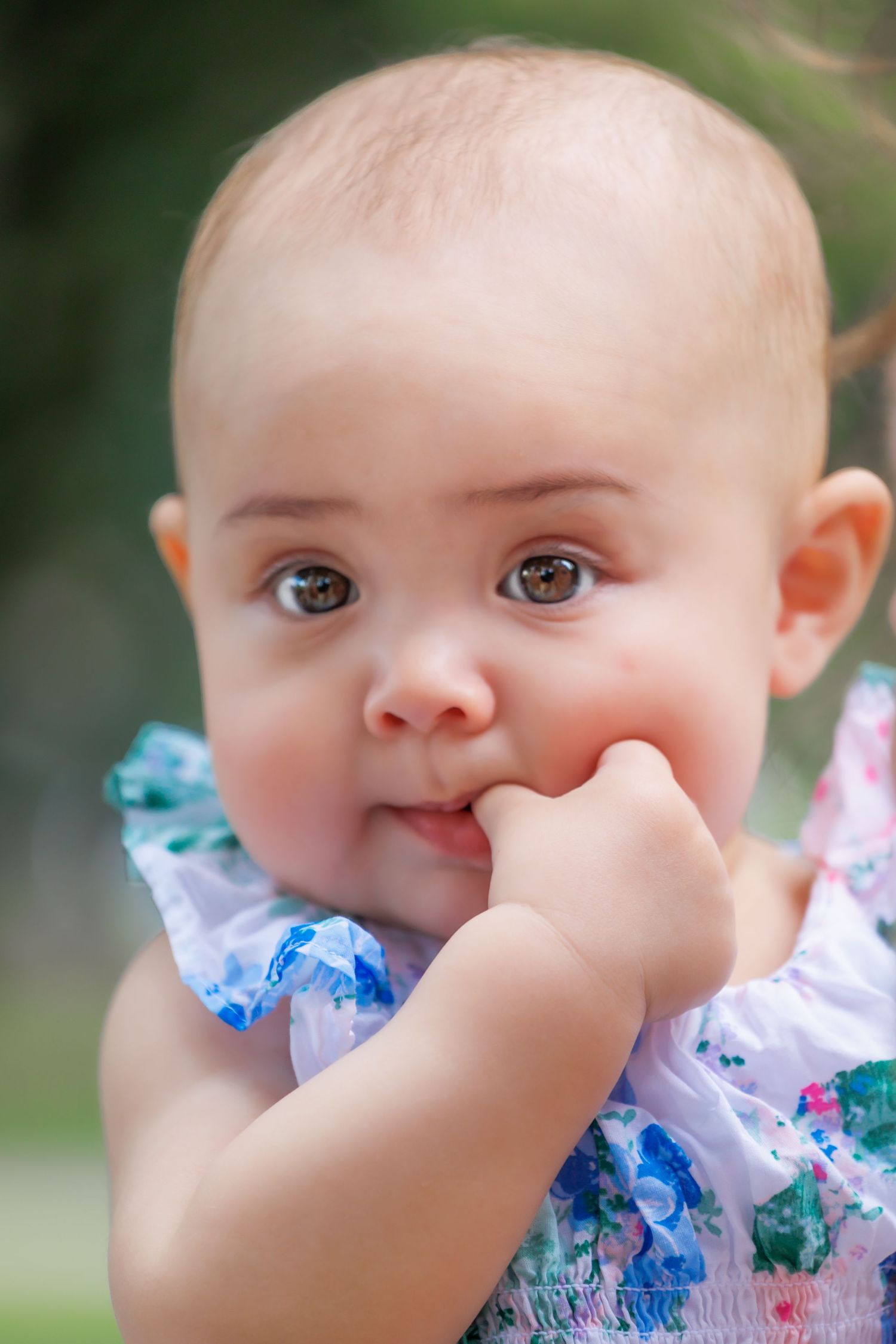 A baby in a floral outfit with their finger in their mouth.