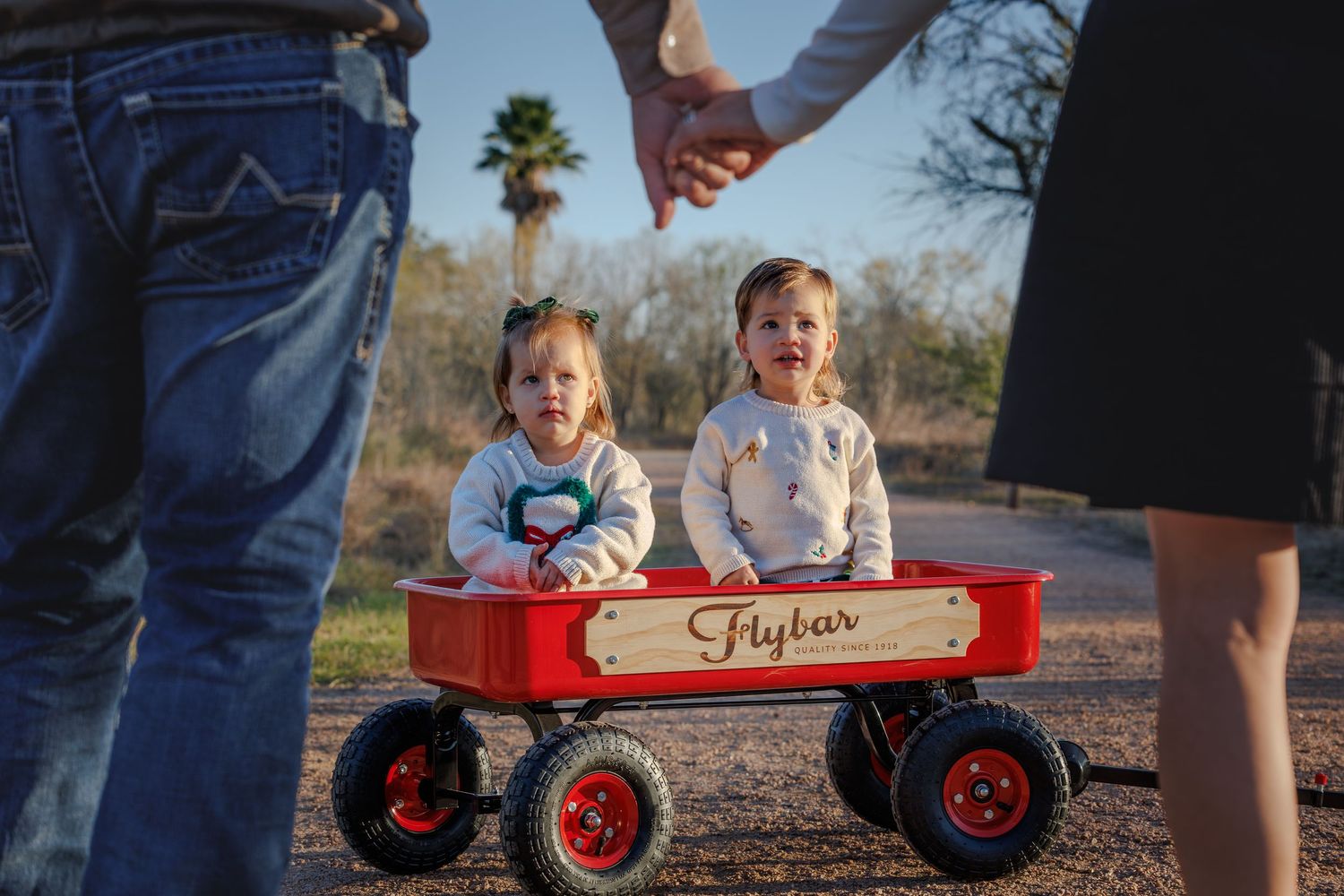 Children sit in a red wagon outdoors while being pulled along a paved path on a sunny day.