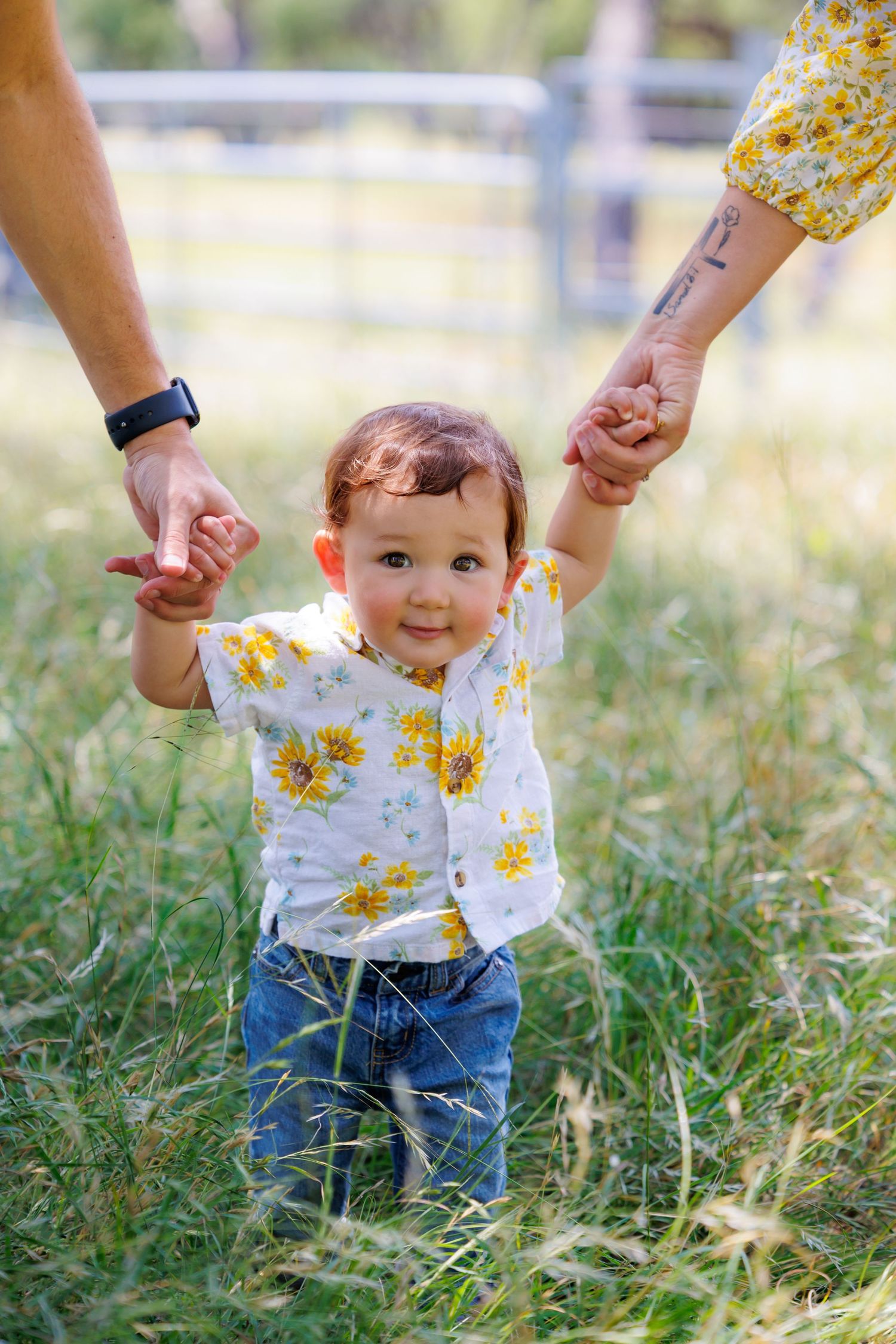 A happy toddler in a floral outfit holds hands with adults while standing in tall grass on a sunny day.