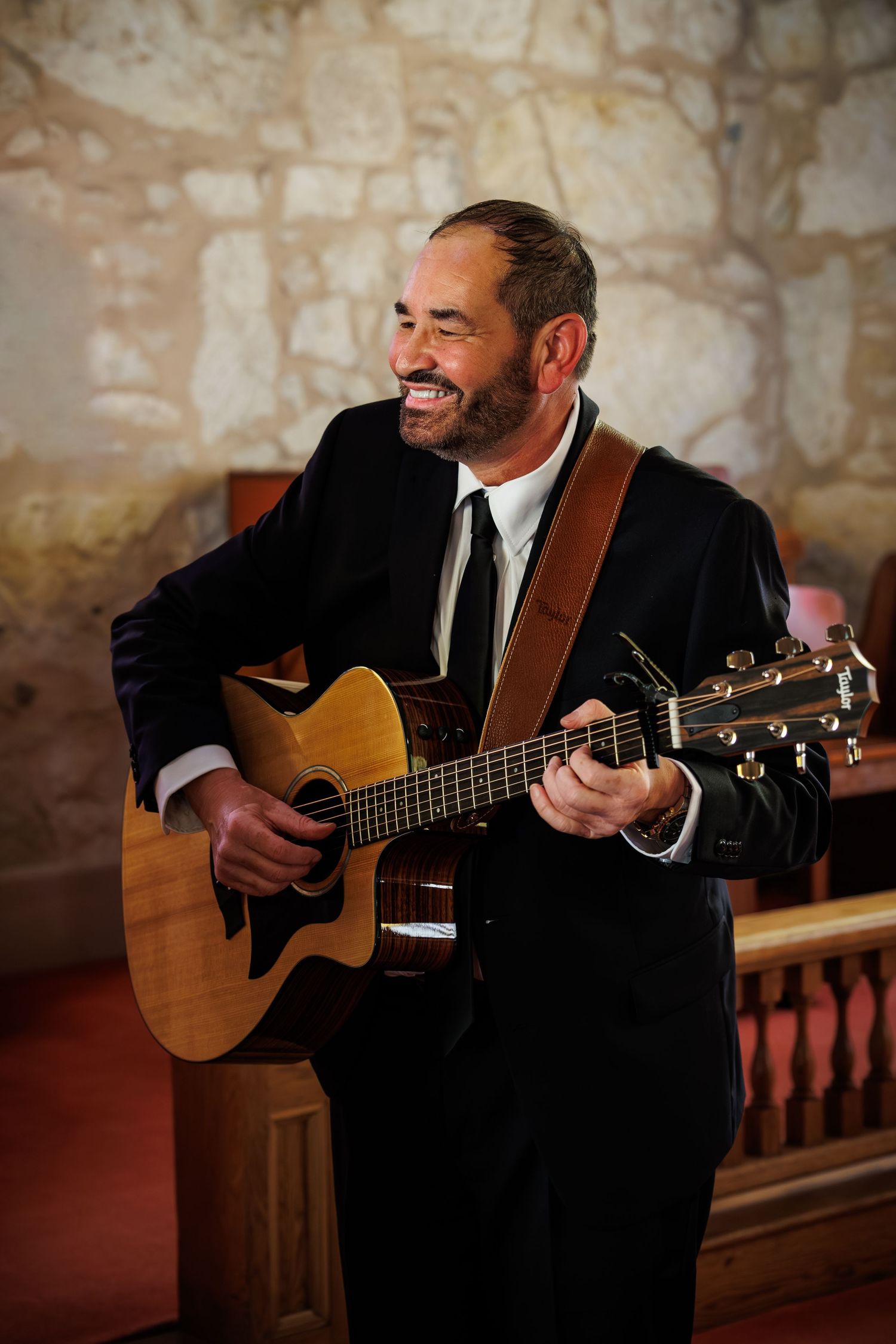 A musician in a black suit plays an acoustic guitar while performing in a rustic stone-walled venue.