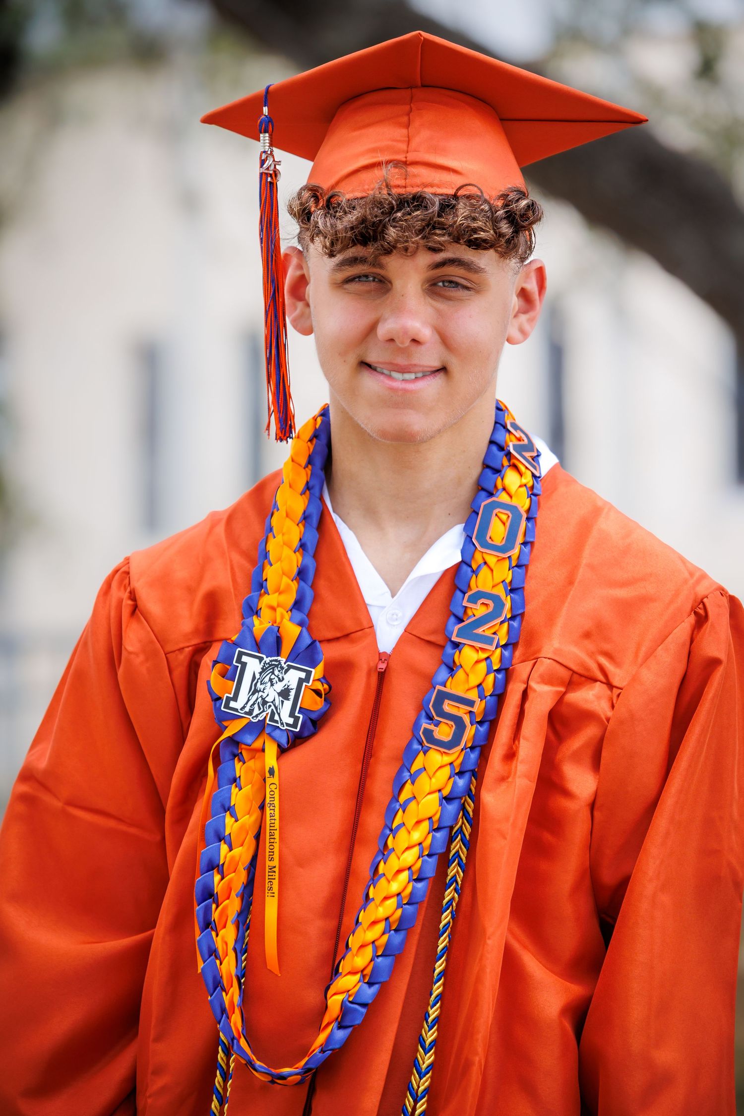 A graduate wearing an orange cap and gown with blue and gold honor cords smiles for a portrait.