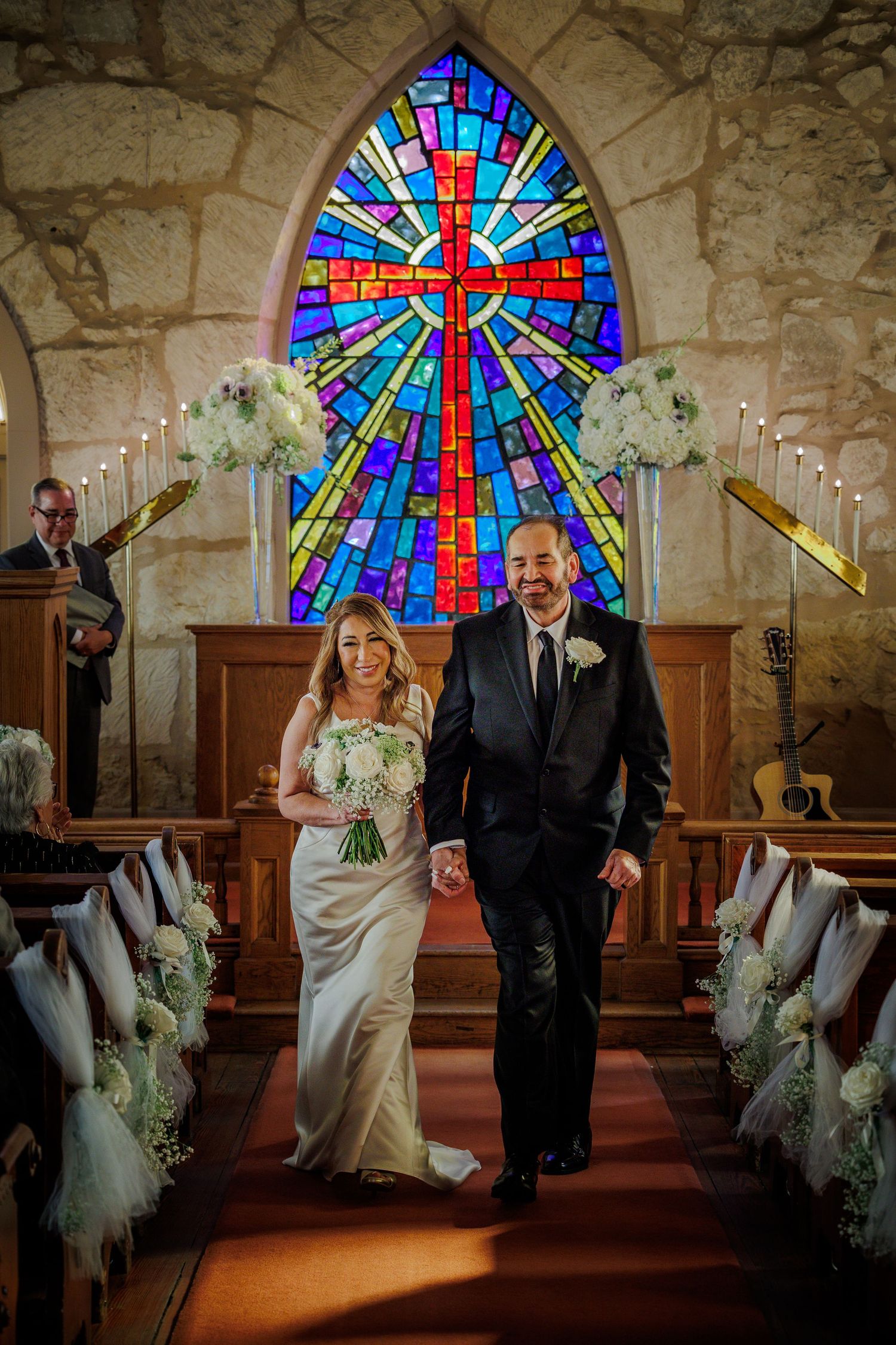 Wedding couple walks down church aisle with colorful stained glass window and stone walls in background.