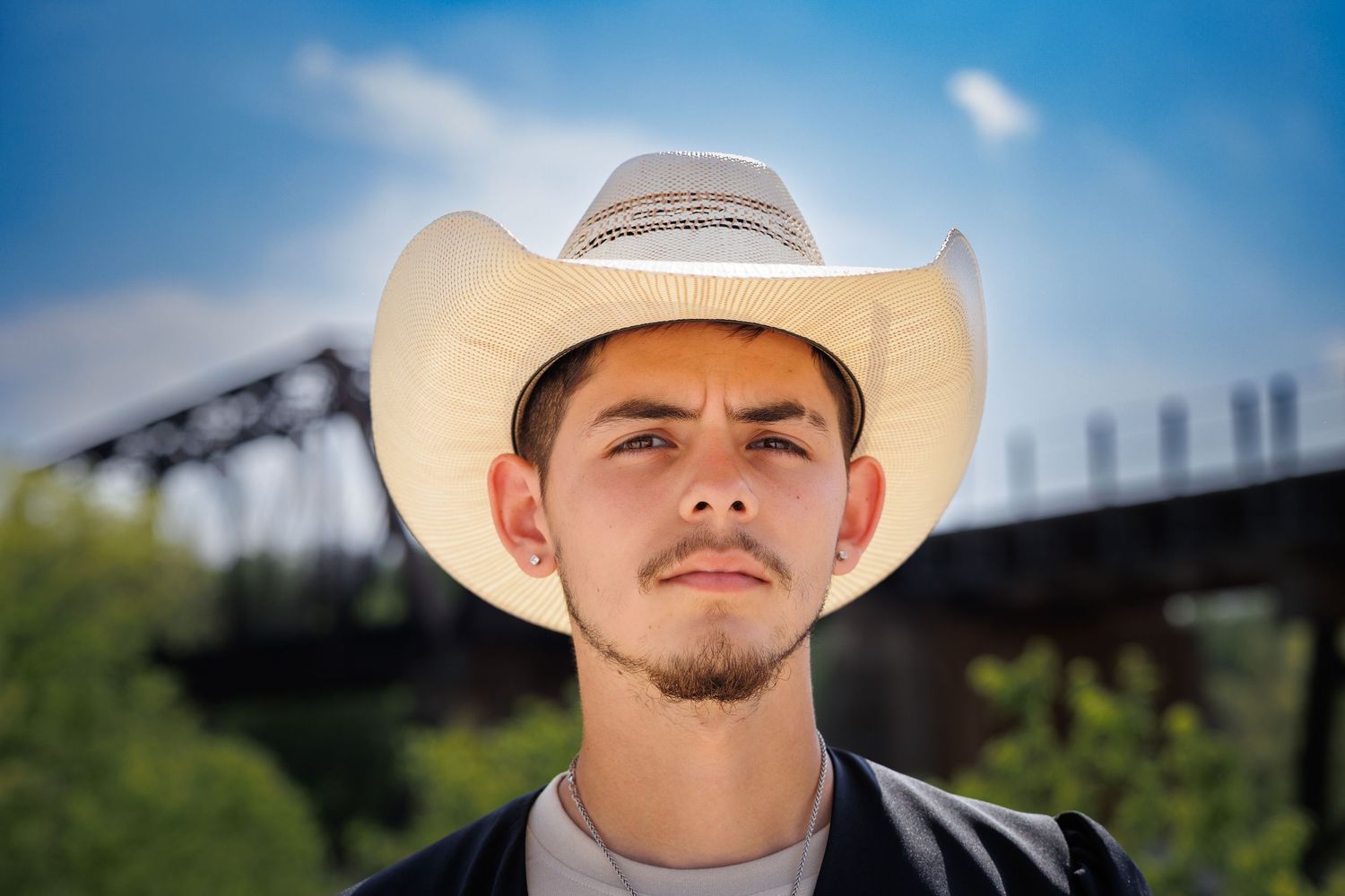 Portrait of young man in cowboy hat with bridge in background under blue sky.