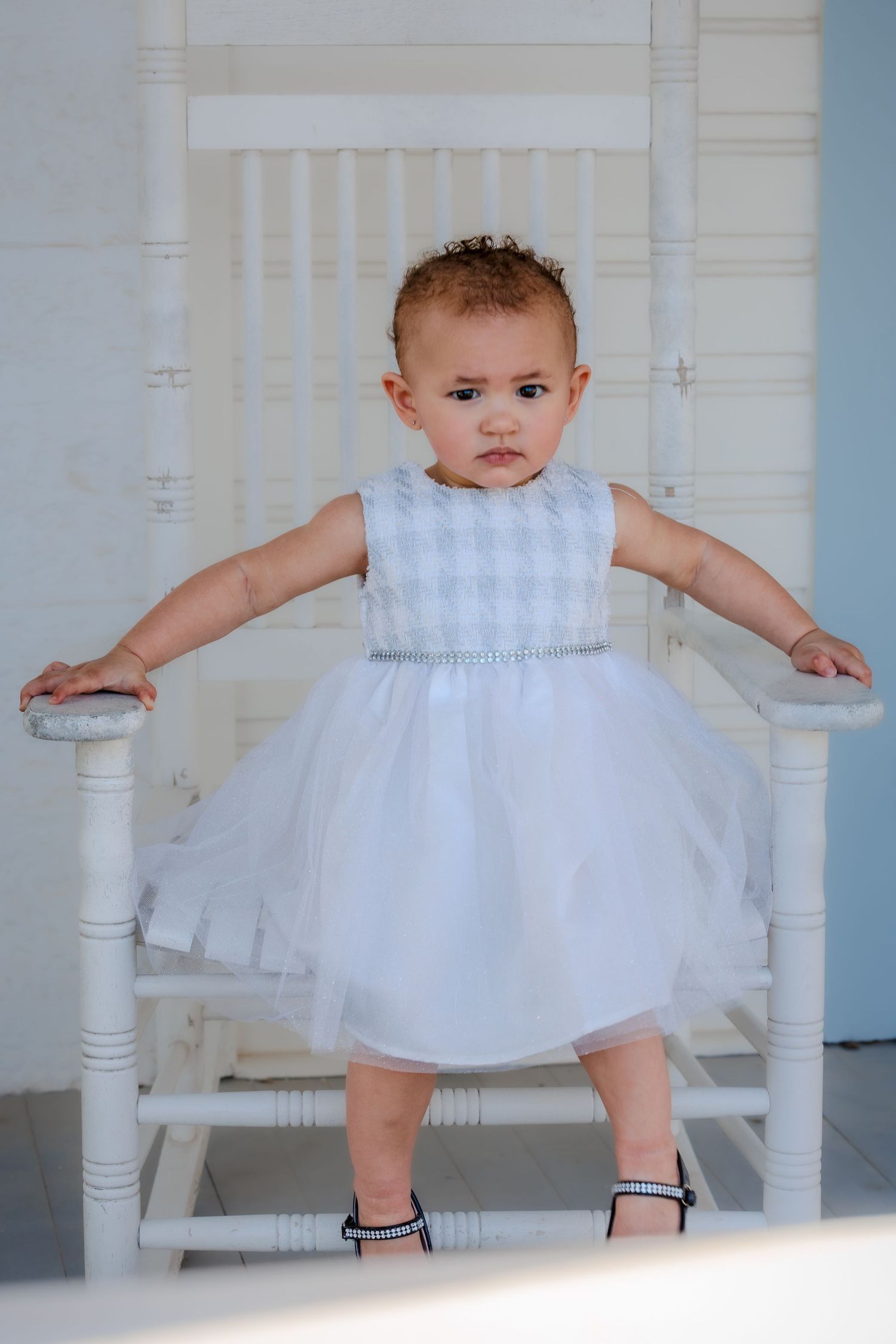 A baby in a white dress and black shoes stands between white porch posts in a series of candid poses.