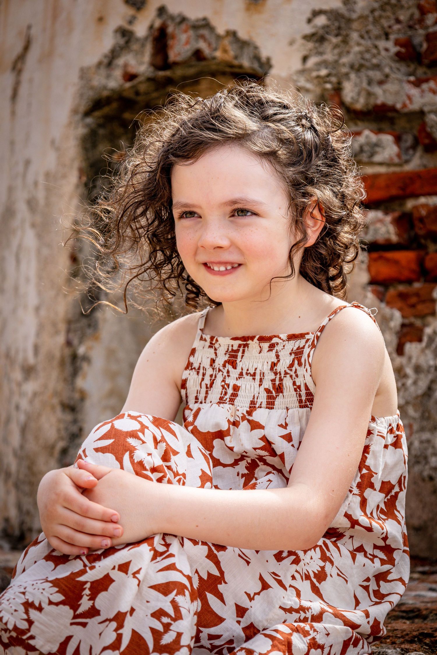 Child with curly hair in floral dress sitting against rustic wall background.