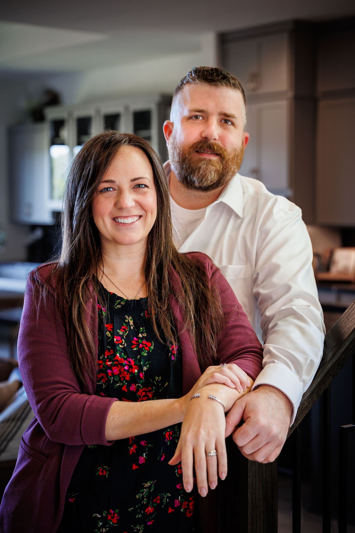A couple stands together in their kitchen, with her wearing a burgundy cardigan and floral top while he wears a white shirt.