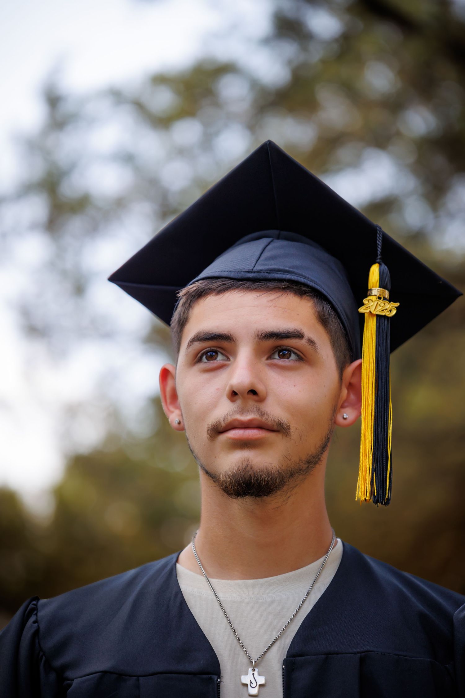 Graduate in black cap and gown with gold tassel looking upward against blurred background.