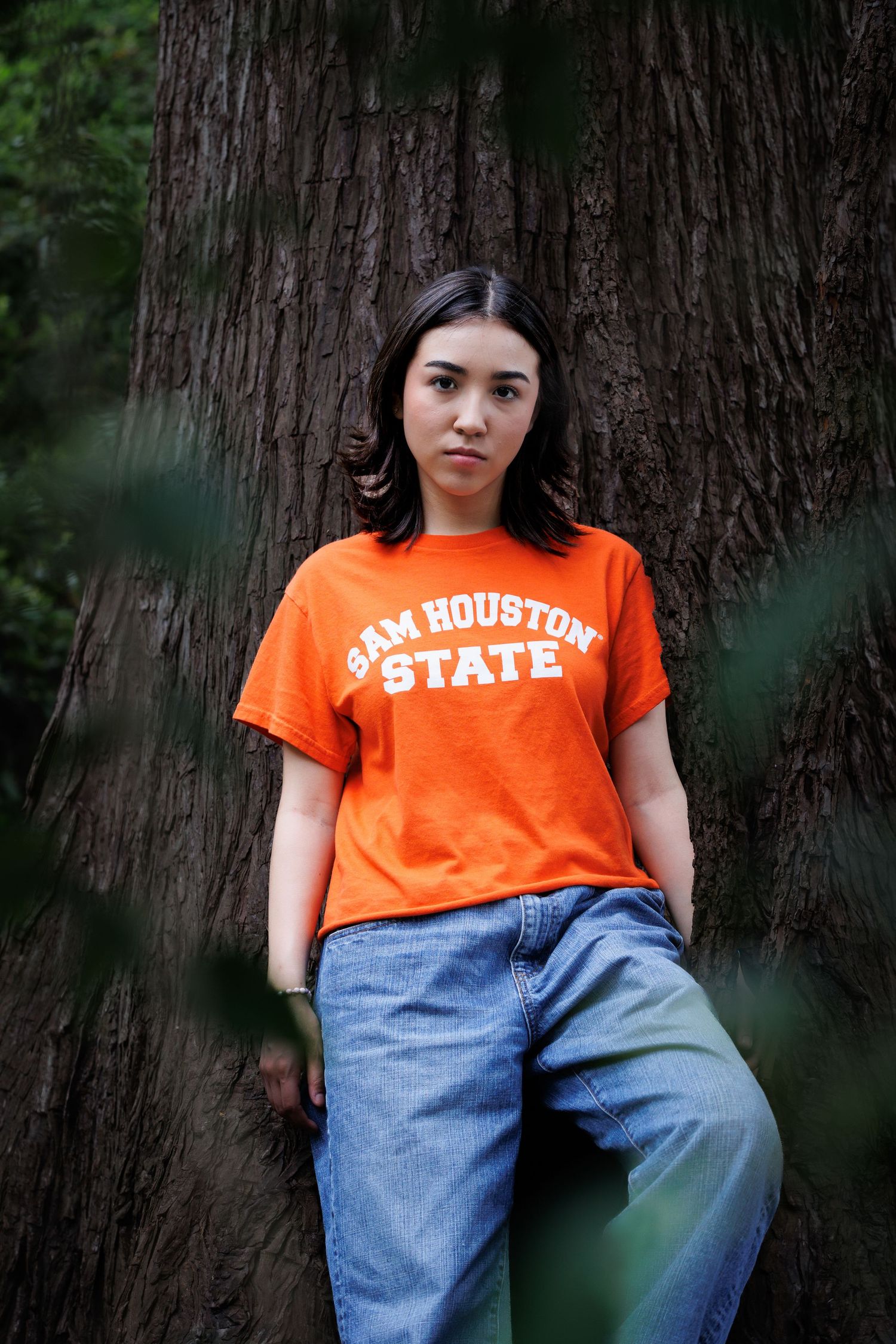 Student wearing orange Sam Houston State shirt leaning against large tree trunk.