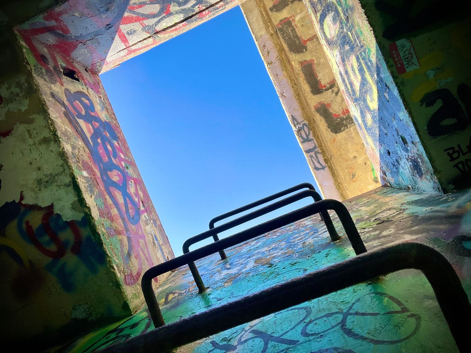 Graffiti-covered interior looking up at blue sky through opening with metal bars silhouetted in foreground.