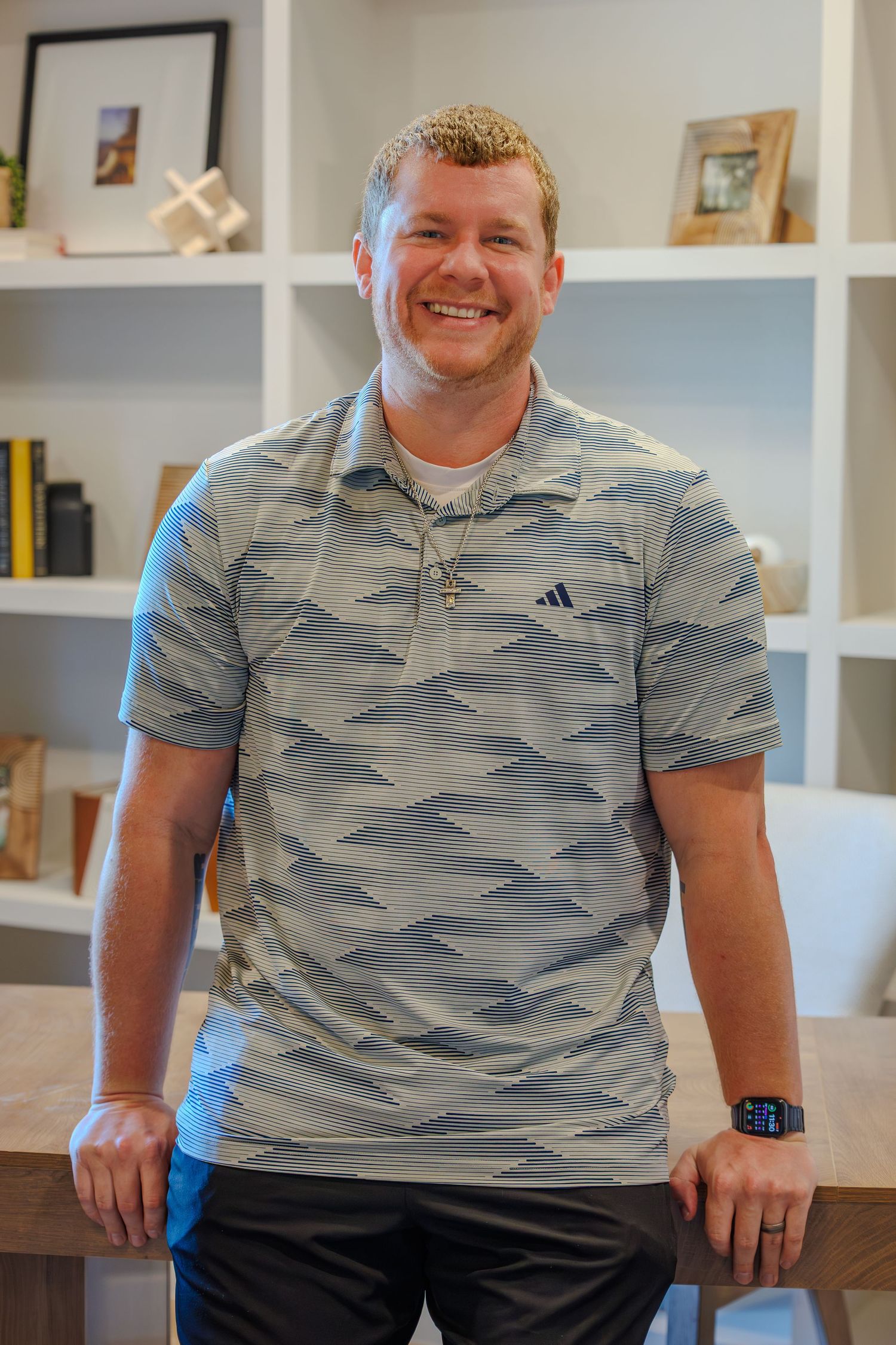 Someone wearing a gray camouflage pattern polo shirt sits and smiles in a room with white shelves in the background.