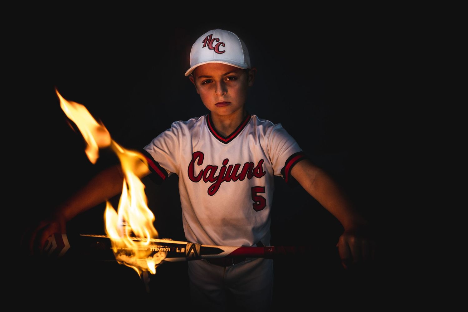 Baseball player in a white Cajuns uniform holds a flaming bat in a dramatic dark setting.