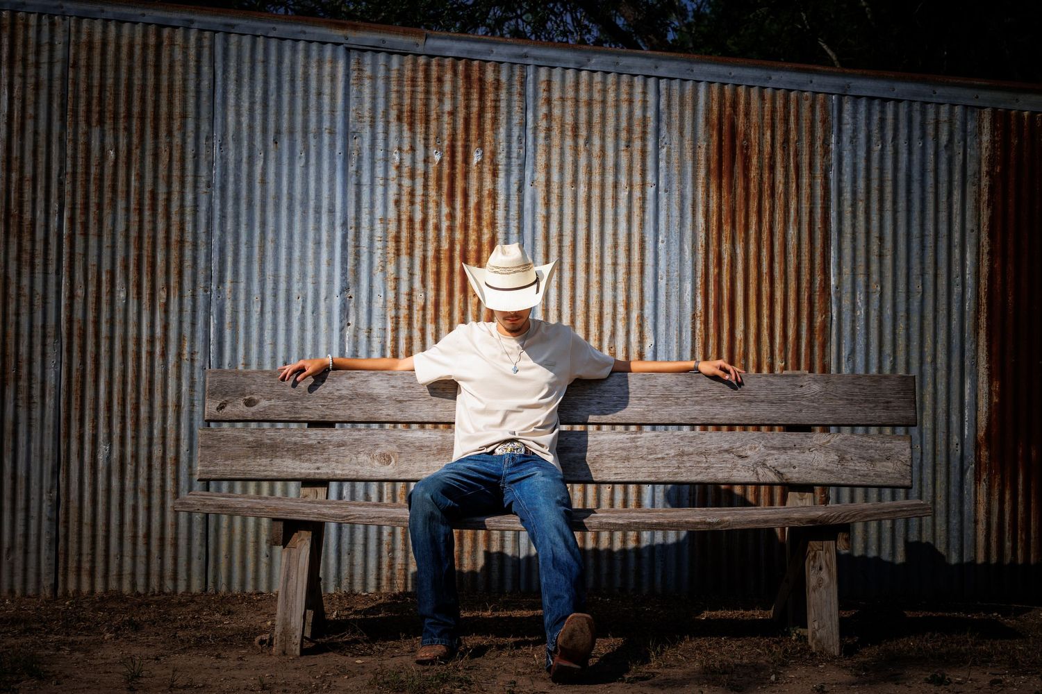 Person in cowboy hat sitting on wooden bench against rustic metal wall.