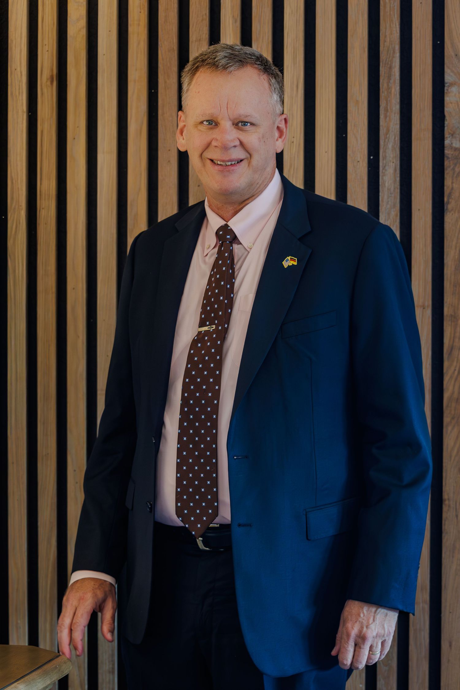 Professional portrait of someone in a navy suit and polka dot tie standing against a wooden slat wall background.