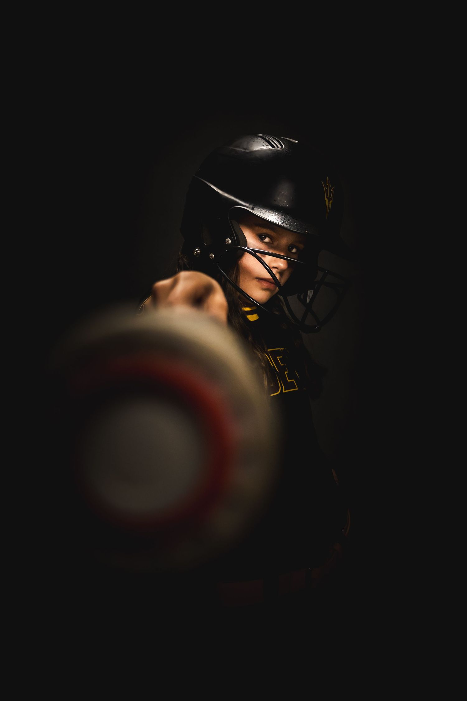 Dramatic portrait of a softball player wearing a black helmet emerging from shadows with moody lighting.