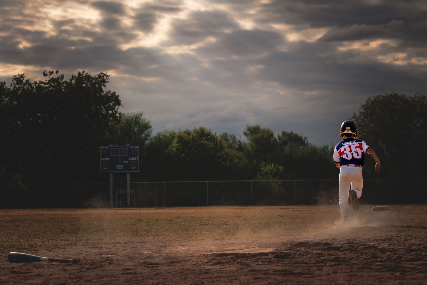 Dramatic storm clouds gather over a dirt baseball field at dusk with trees silhouetted against the glowing sky.