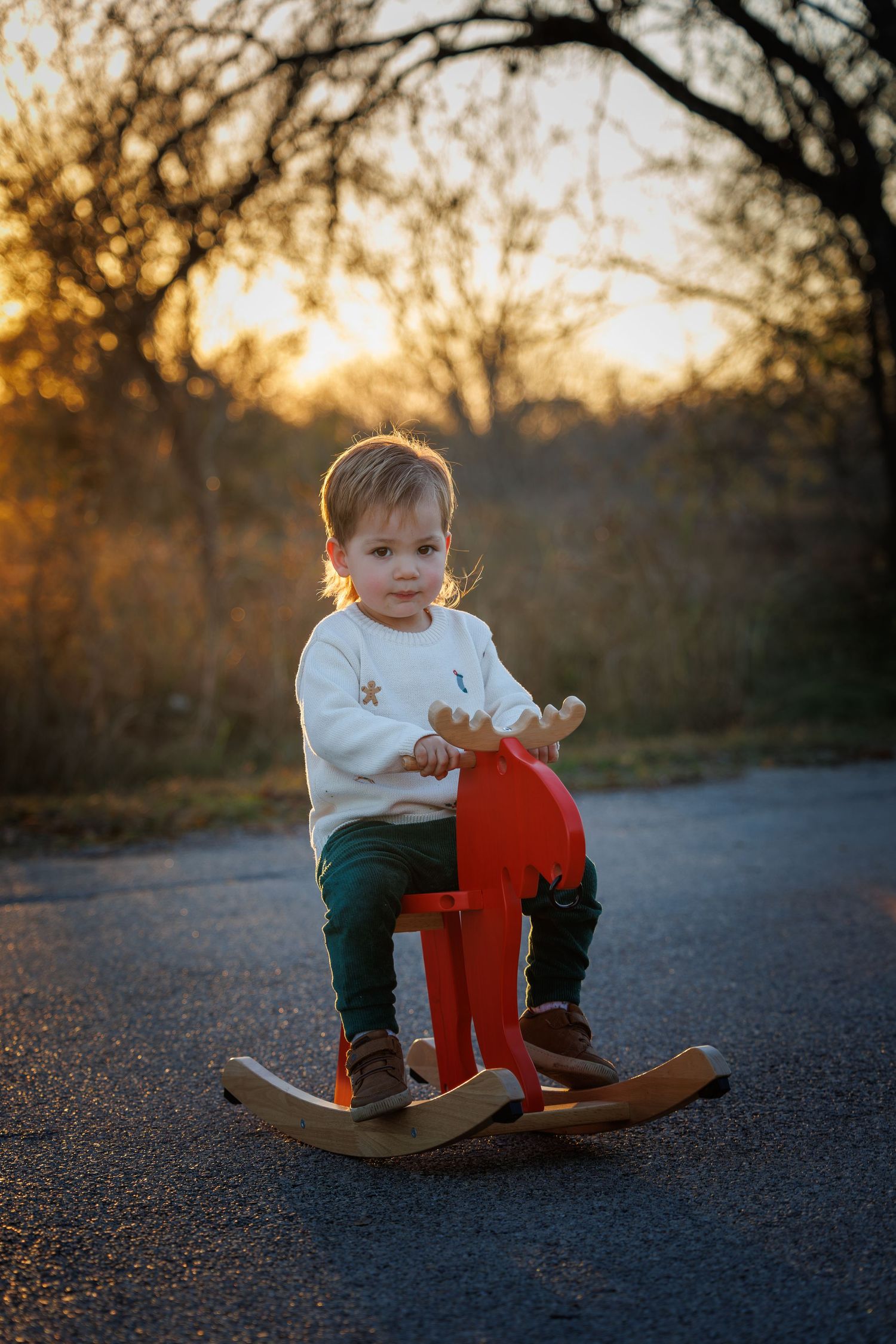 A small child rides a red rocking horse on a path during golden sunset with silhouetted trees in background.