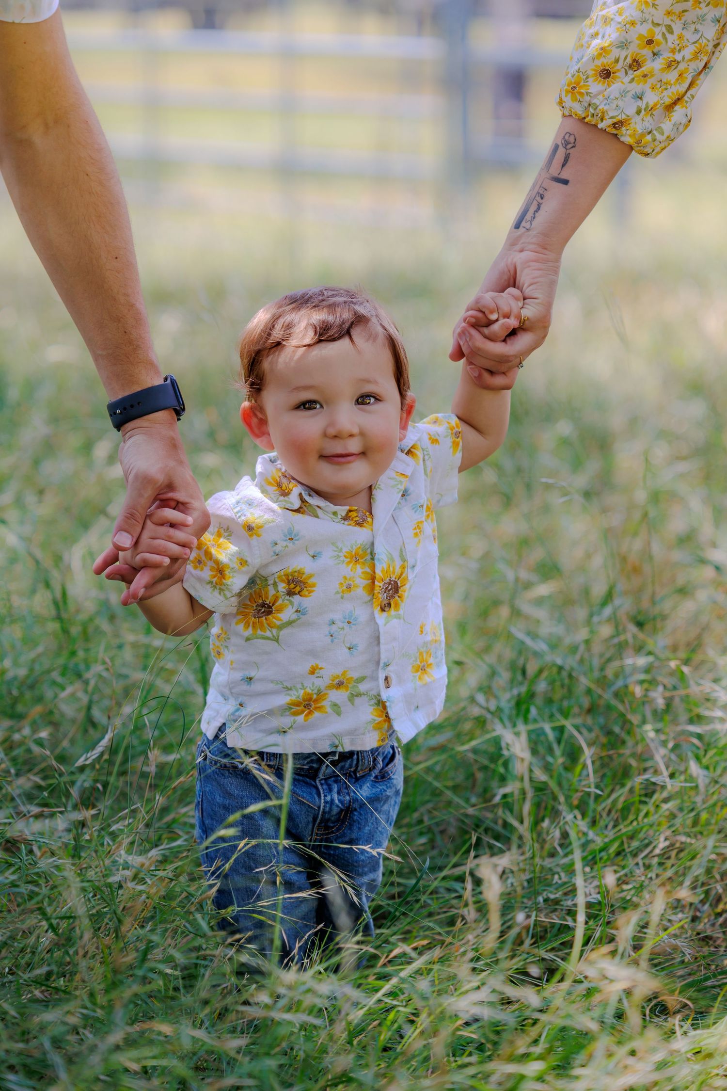 Small child in a floral shirt walking supported by two adult hands in a grassy field.