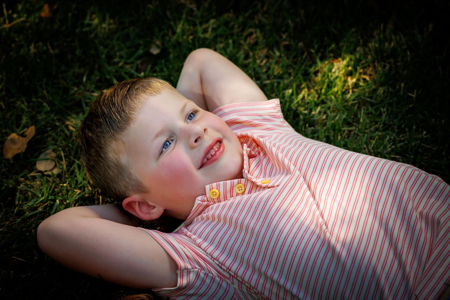 Child in striped shirt lying on grass and laughing in sunlight.