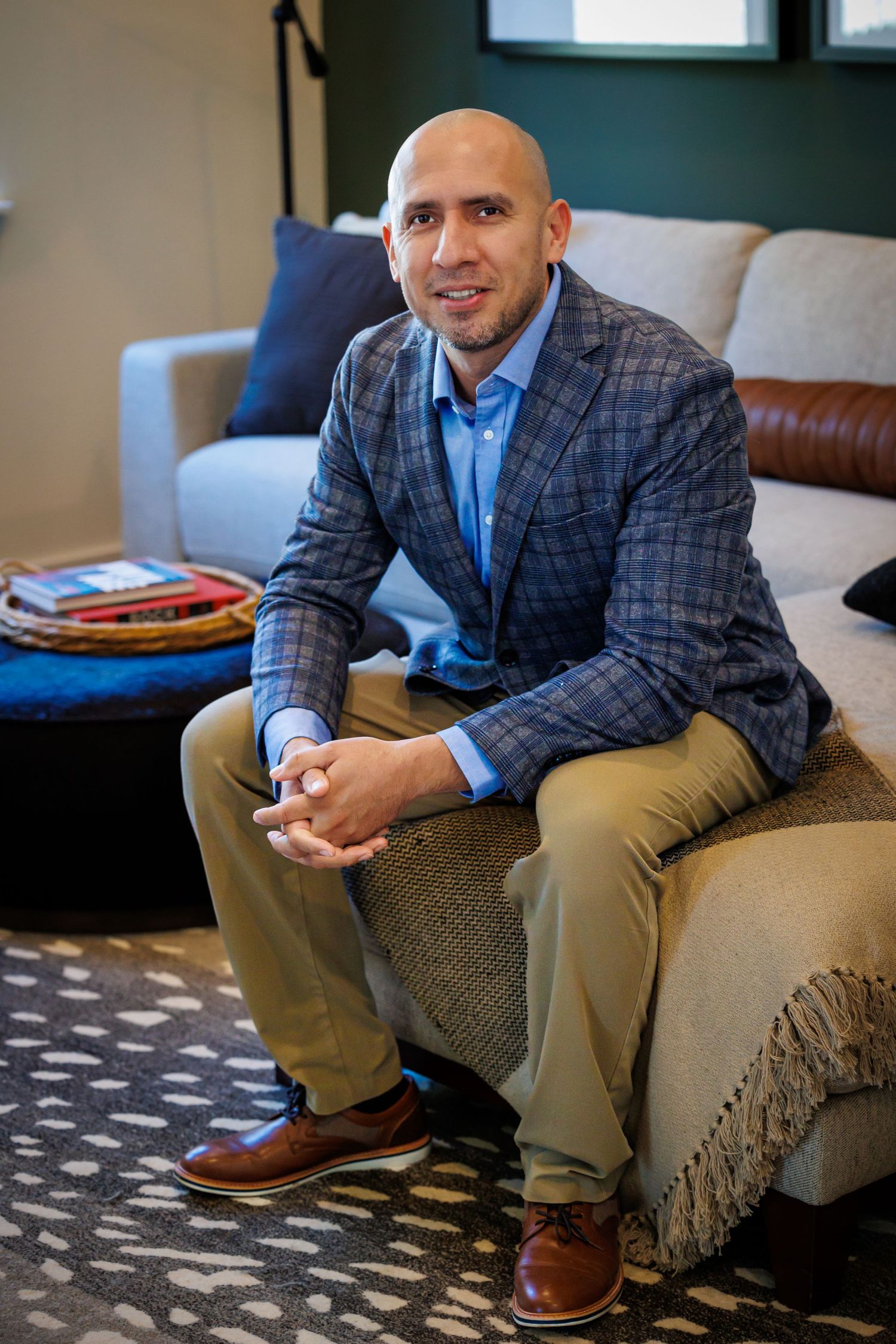 Professional wearing a plaid blazer and blue shirt sits casually on a couch in a modern living room setting.