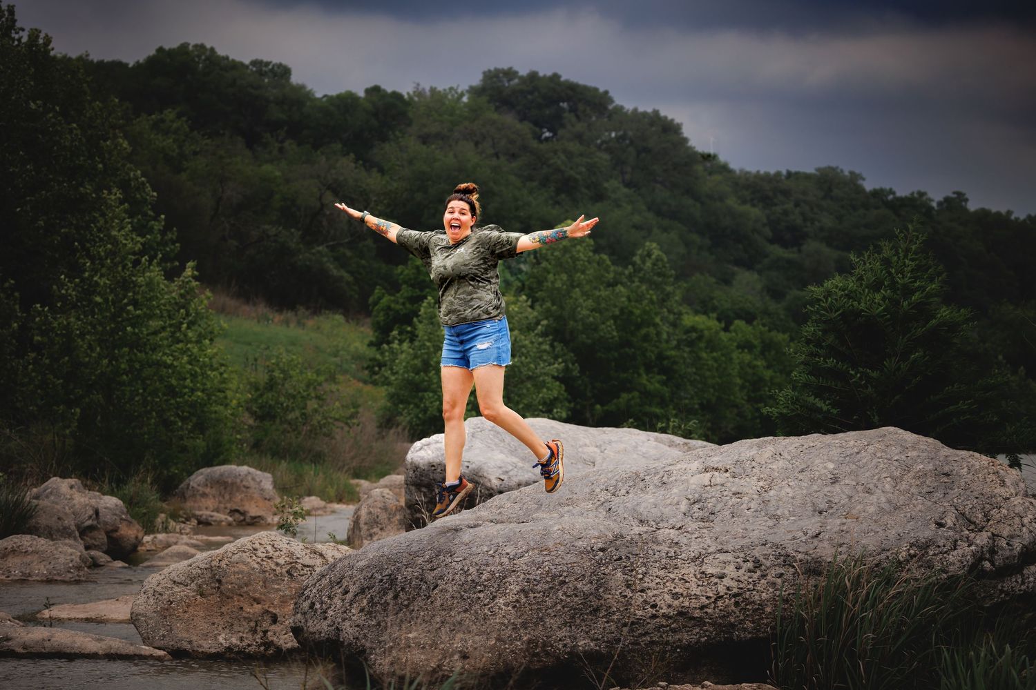 A figure stands with outstretched arms on a large rock formation surrounded by green forest under a moody sky.