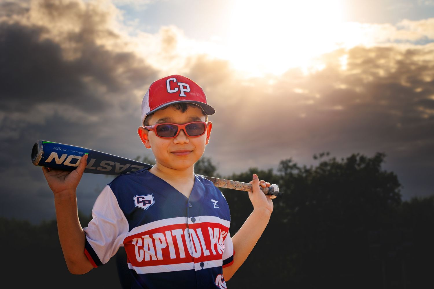 Youth baseball player in Capitol Park uniform posing with bat against dramatic sunset.