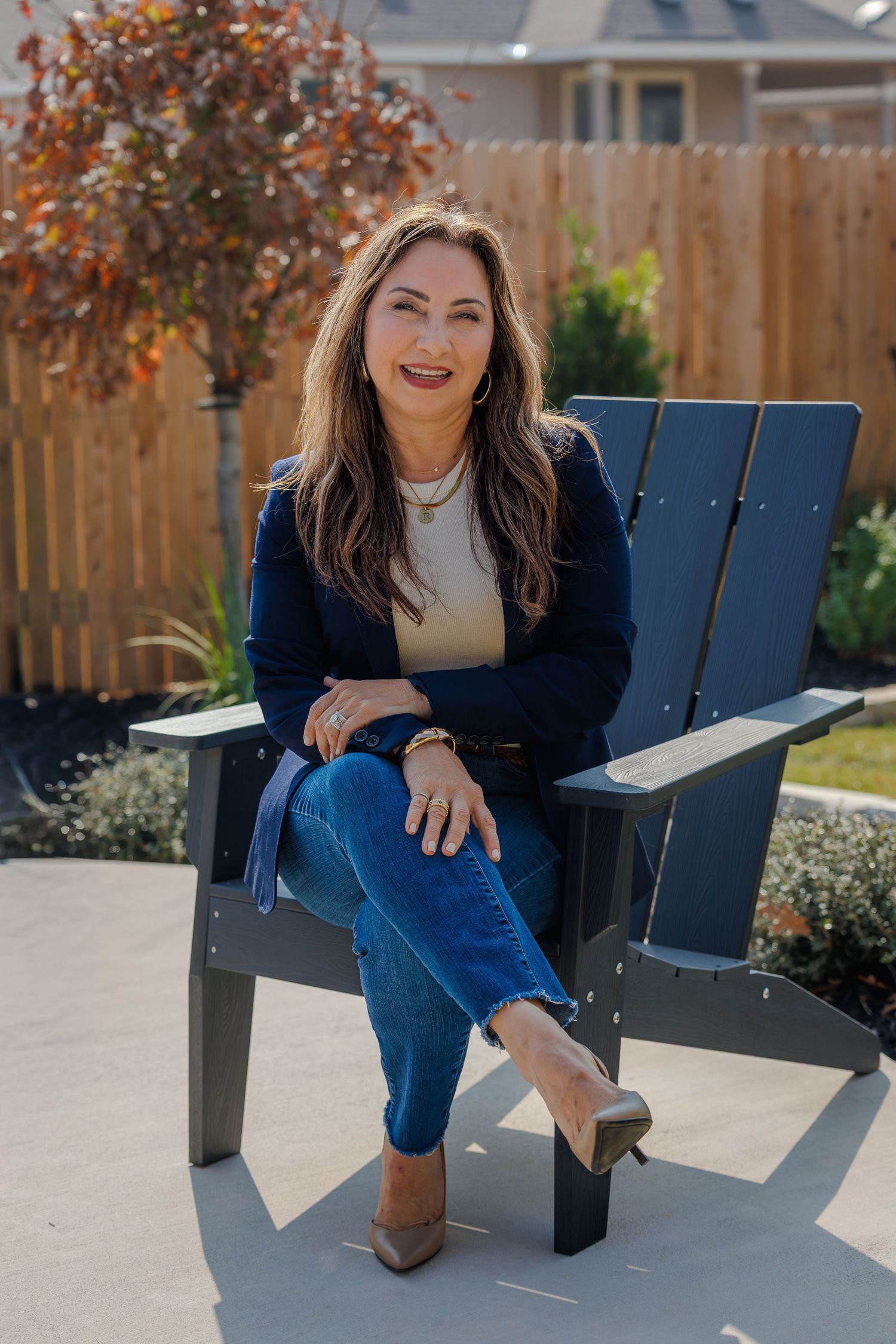 Casual outdoor portrait of someone in a navy blazer and jeans sitting in a dark Adirondack chair on a patio.