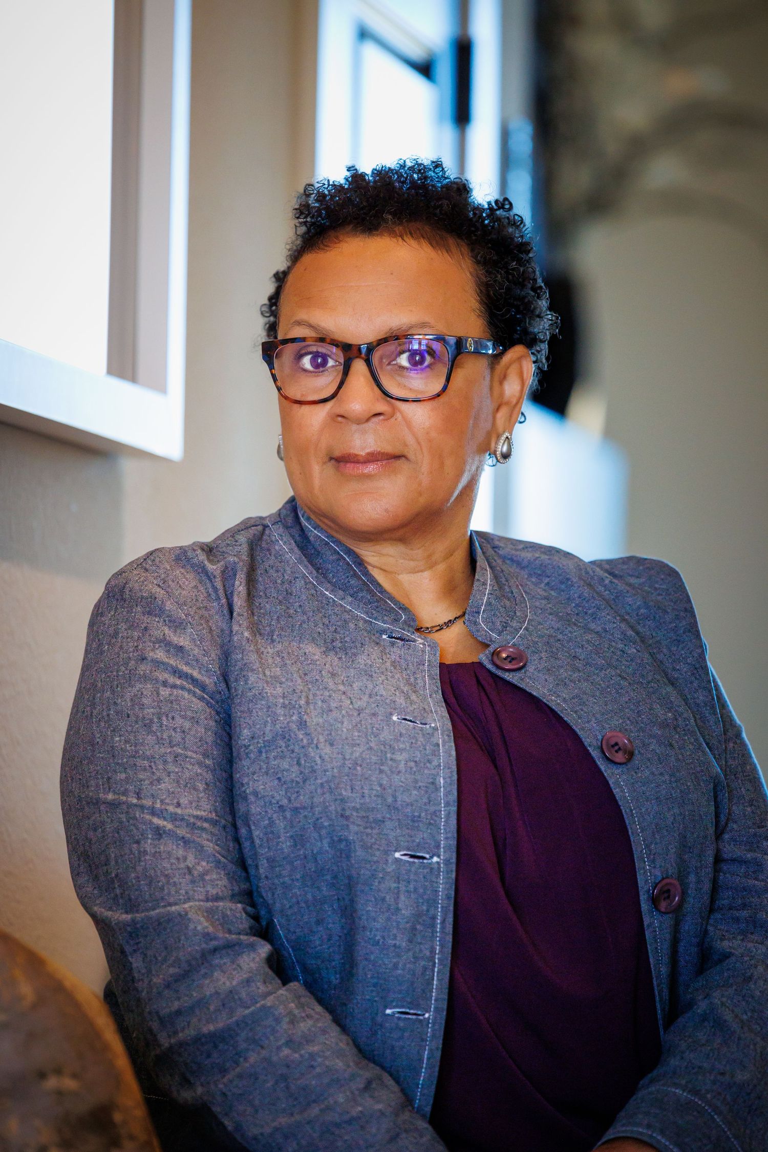 Professional portrait of someone wearing glasses and a grey blazer over a purple top in an indoor setting.
