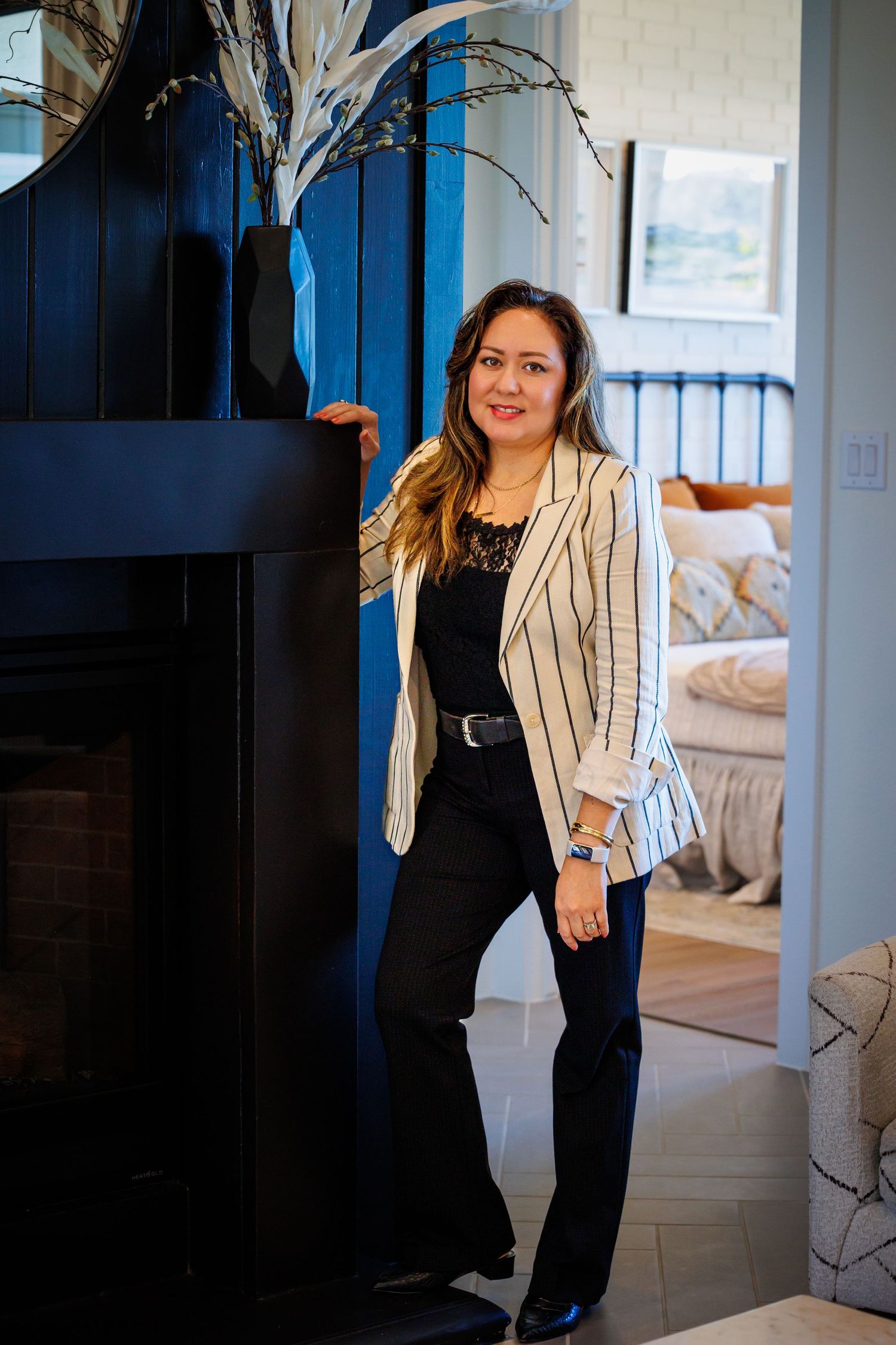 A person wearing a striped blazer and black outfit stands by a blue wall and fireplace in a modern interior setting.