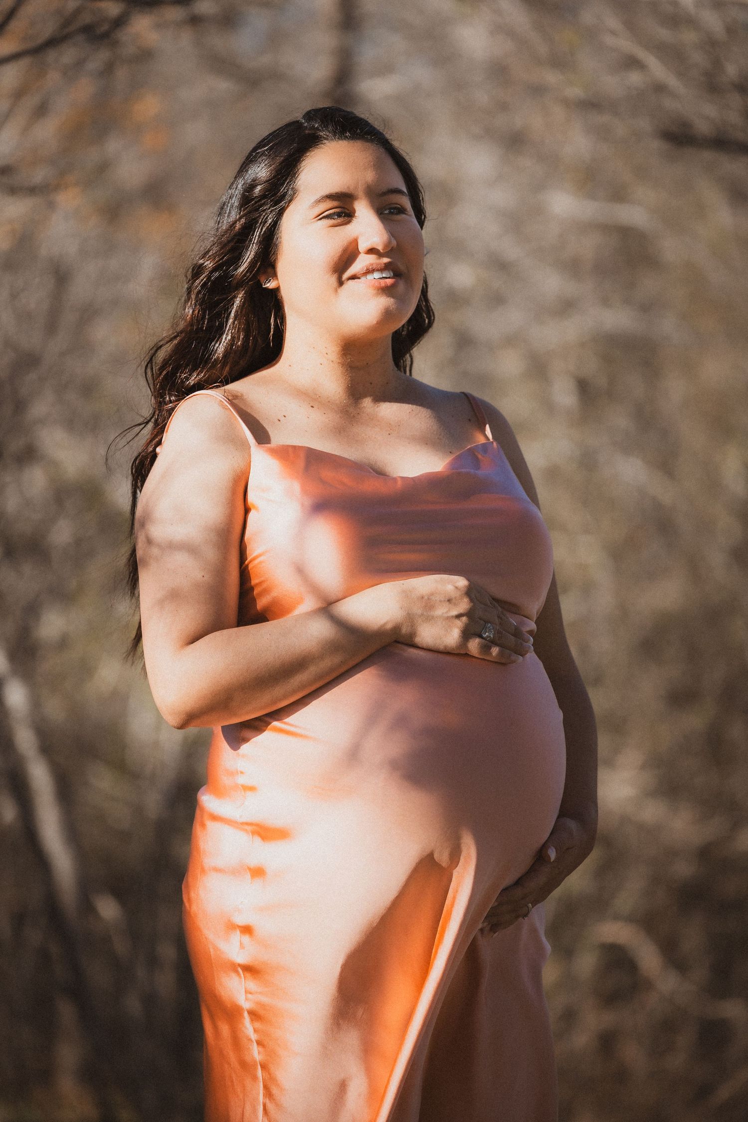 Maternity photo of woman in orange silk dress against natural backdrop.