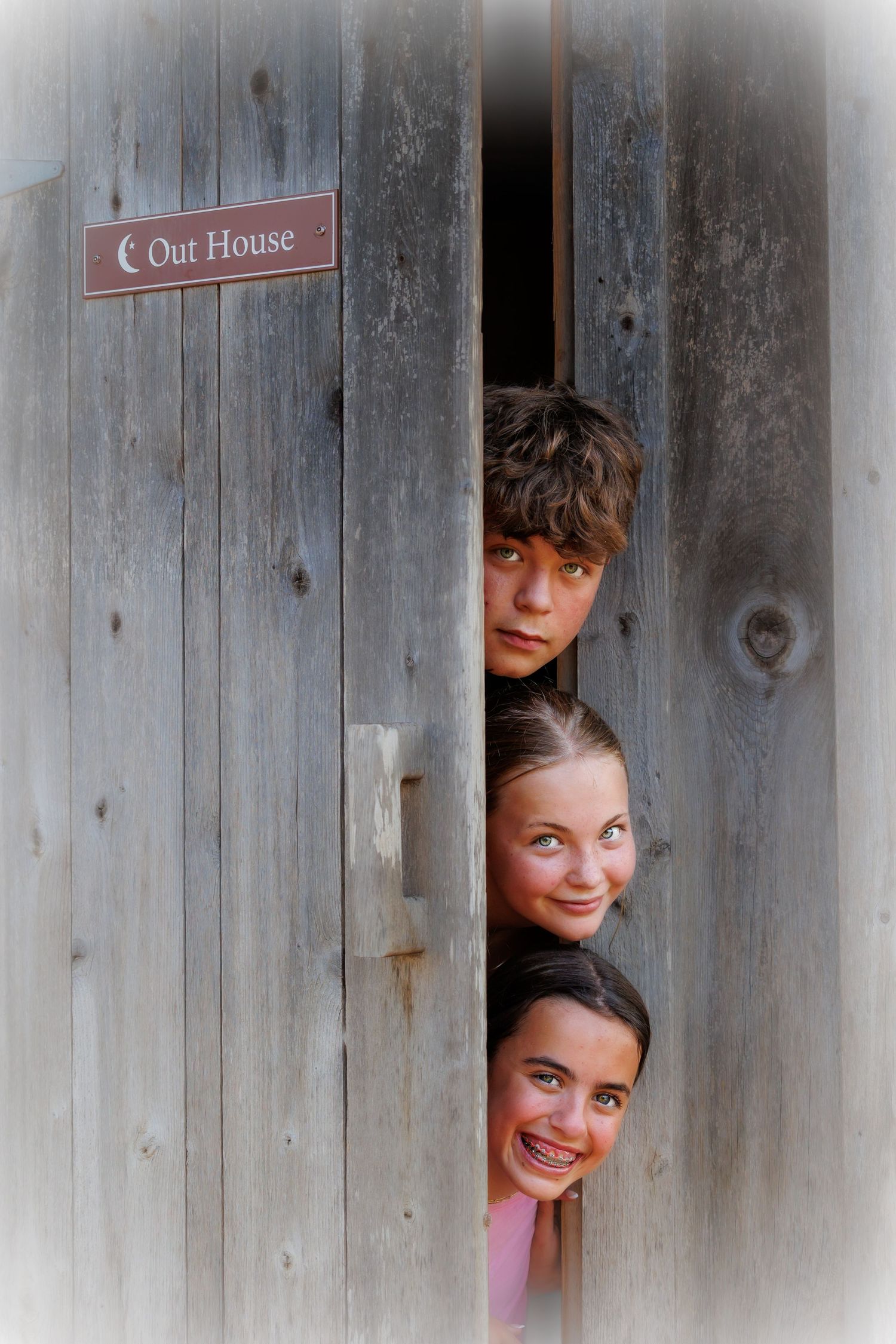 Three people peek playfully through a gap in an old wooden outhouse door.