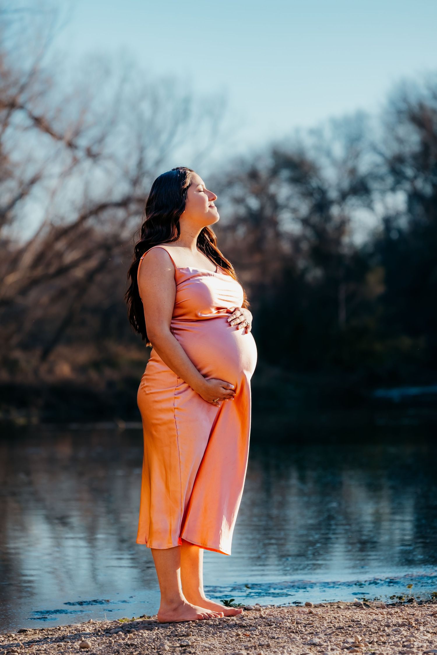 Maternity photo of expecting mother in peach dress standing by lake at sunset.