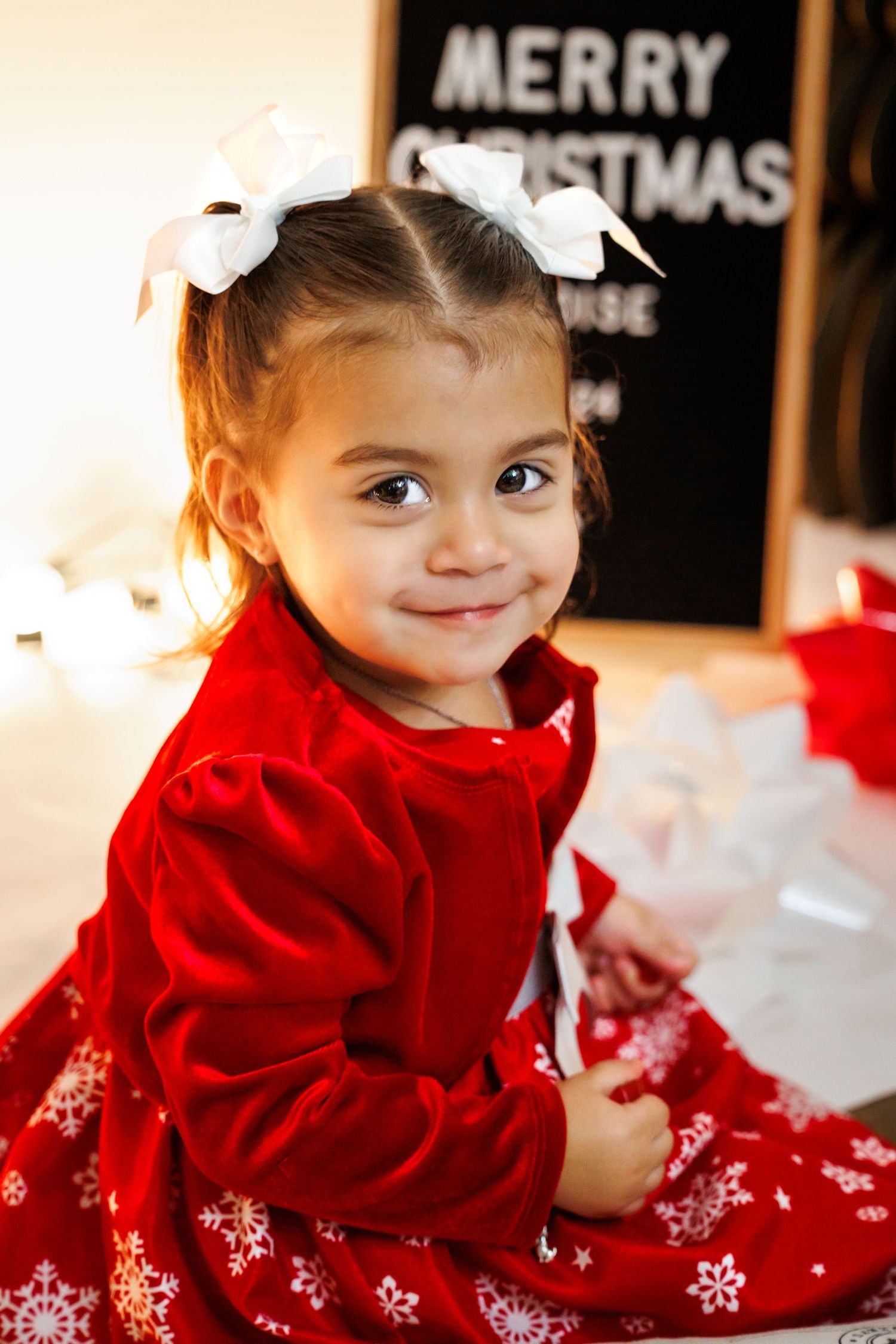 Holiday portrait in red velvet dress with white bows near Christmas lights and decorations.