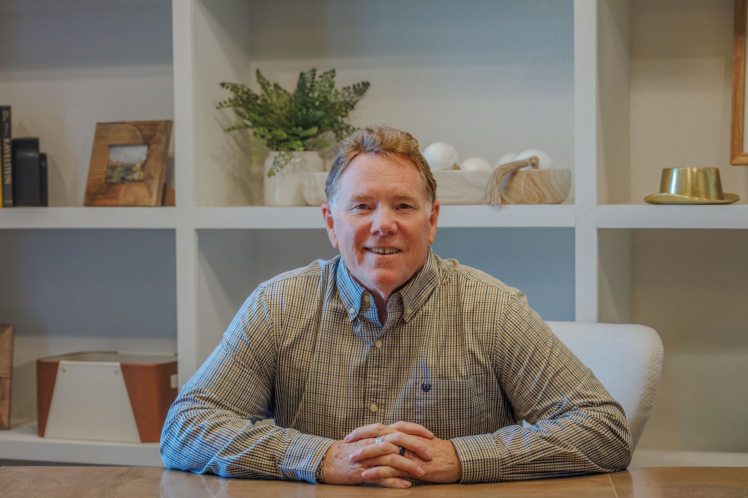 Senior person wearing a plaid shirt sits at a desk with shelving and decor visible in the background.
