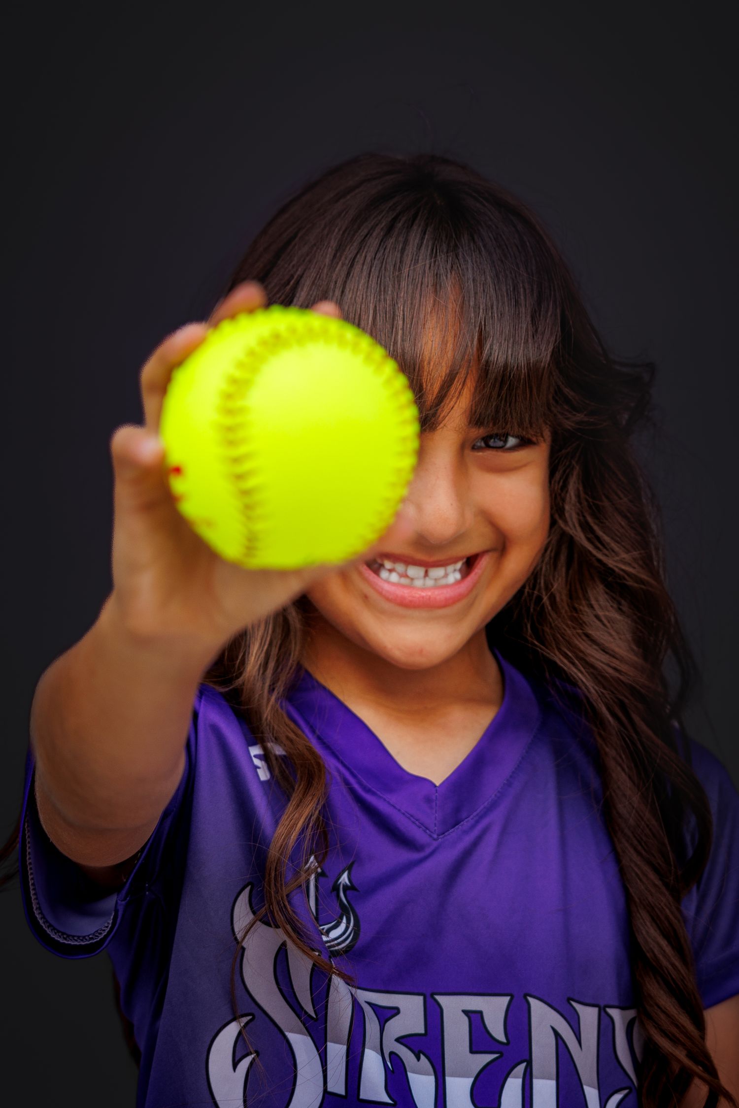 Softball player in purple uniform holds up bright yellow softball with a cheerful smile against dark background.