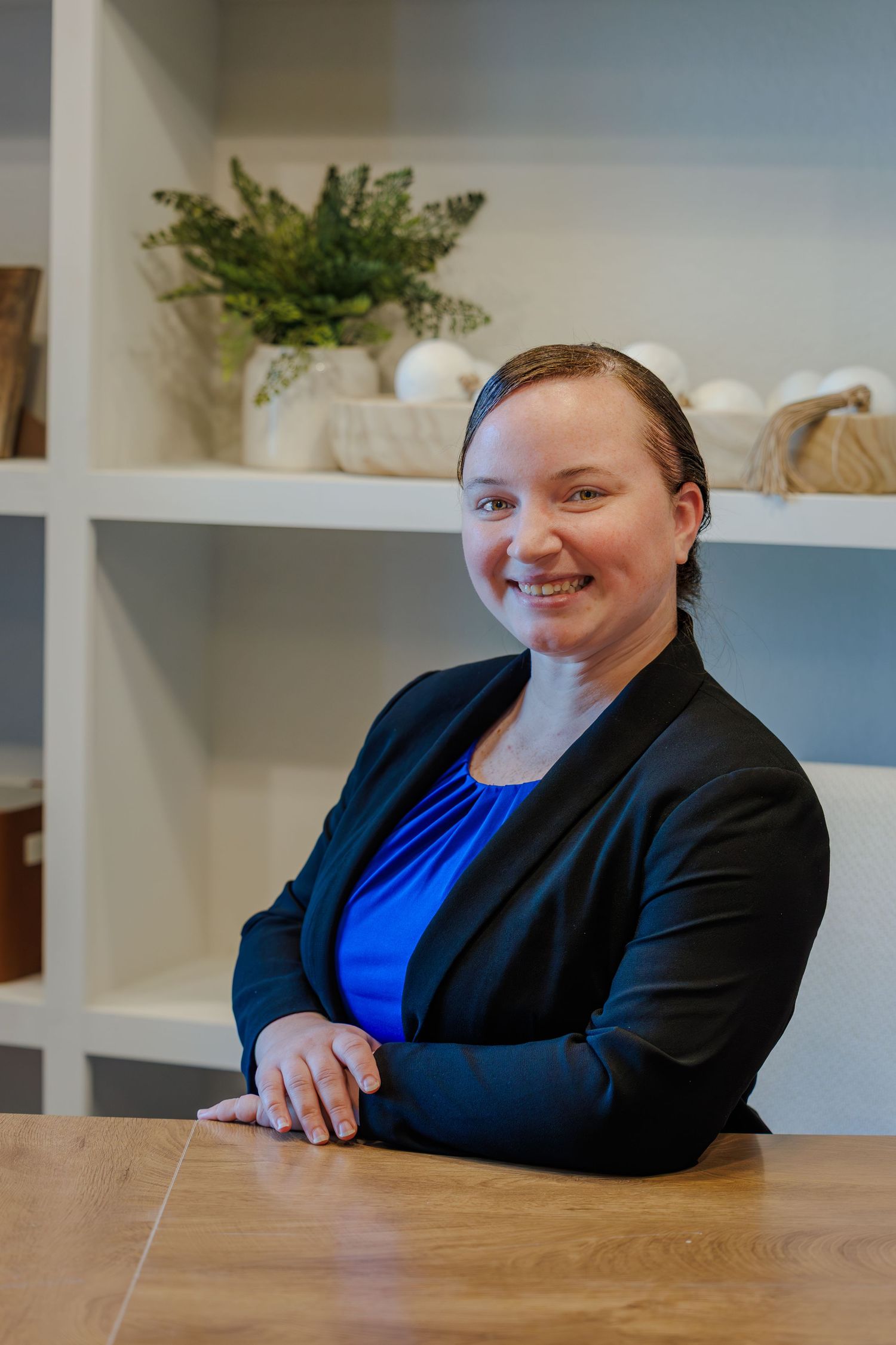 A person in a black blazer and blue top sits at a wooden table while smiling.