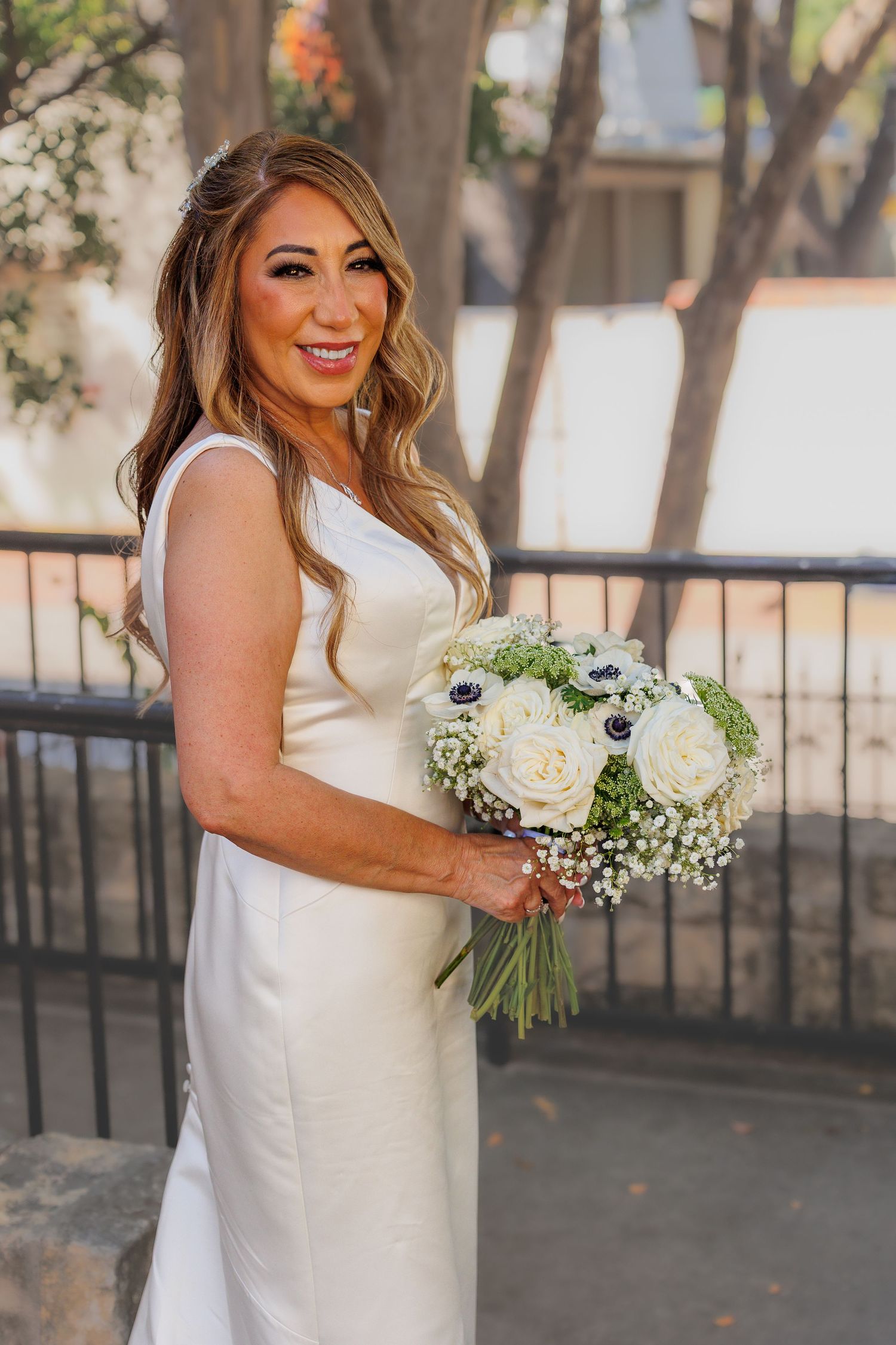 Bride in elegant white sleeveless gown poses outdoors with a beautiful bouquet of white roses and baby's breath.