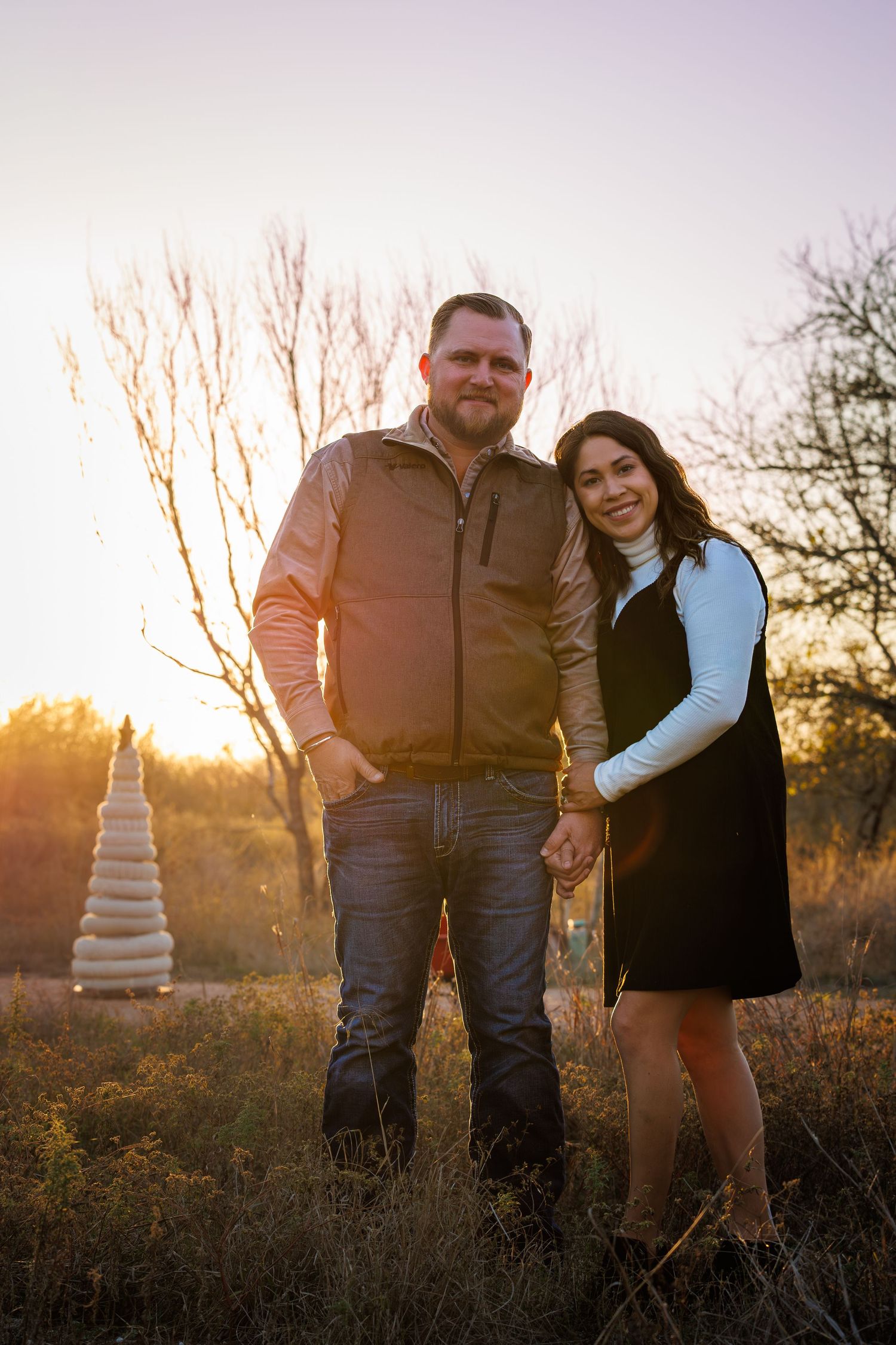 A couple poses together in a desert landscape at sunset with a tiered white cake visible in the background.