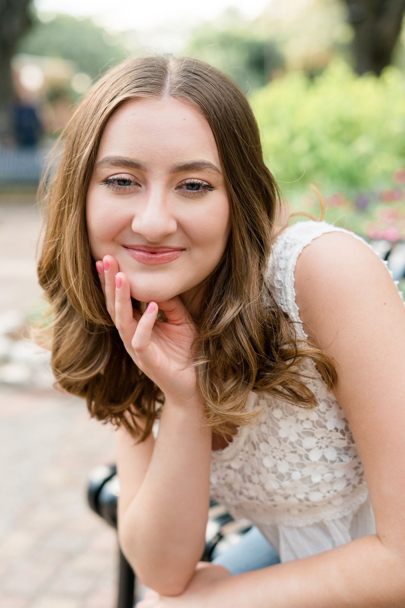 Young woman with wavy brown hair in white lace dress, smiling softly while resting chin on hand outdoors.