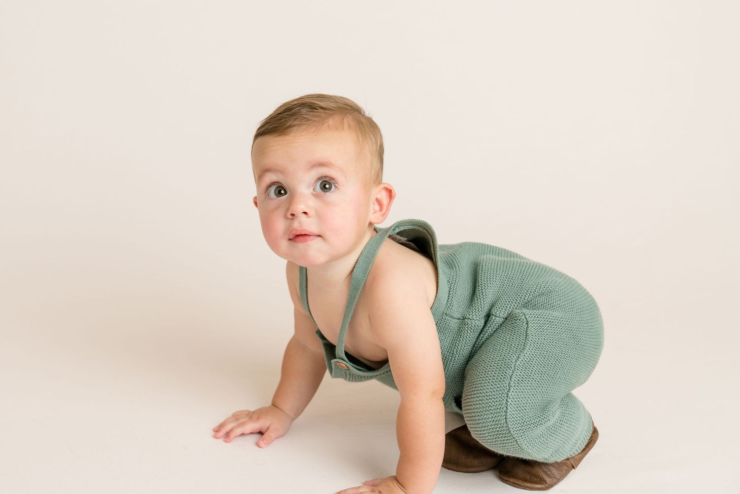 Baby boy in green romper crawling on white studio floor looking at camera.