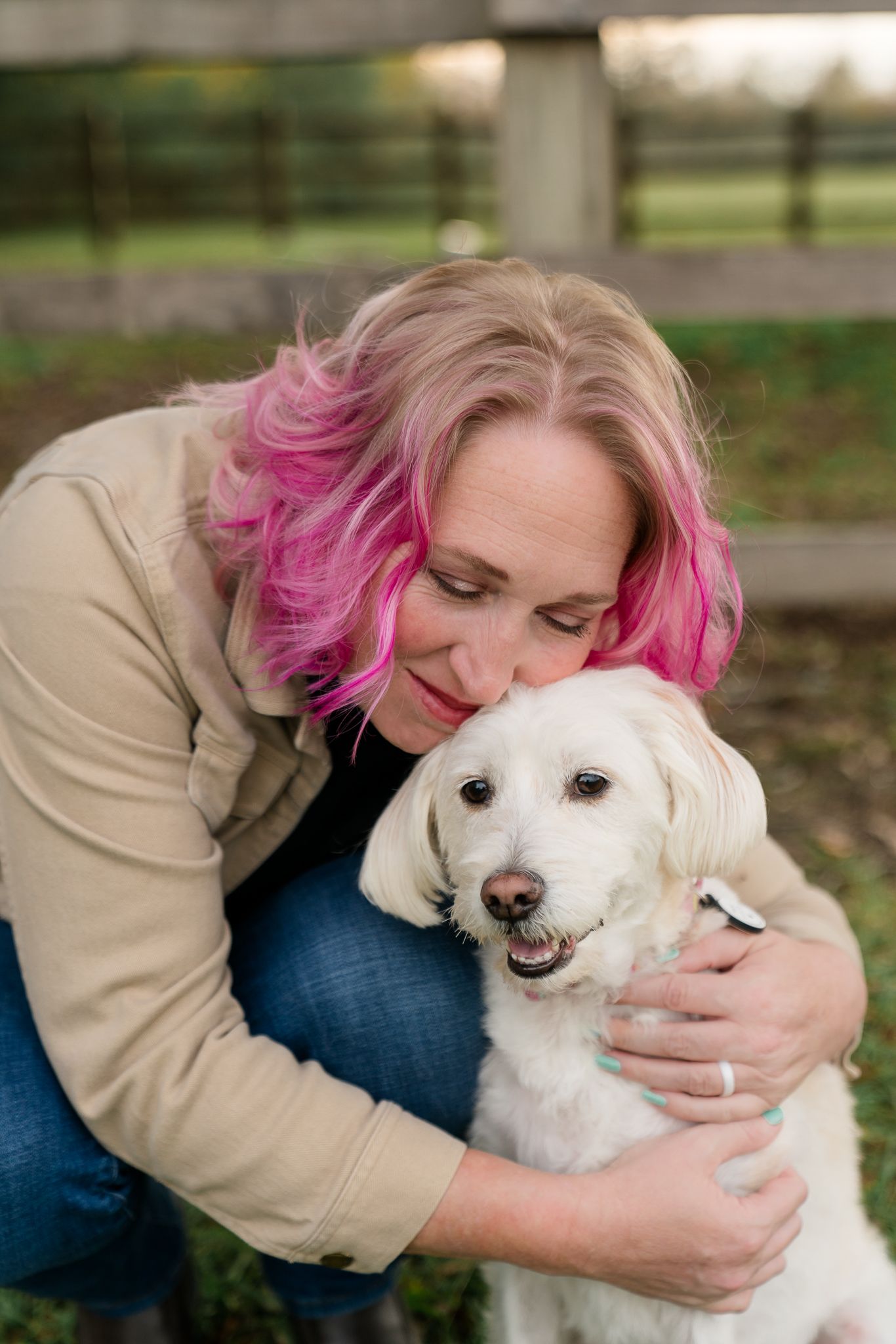 A woman with pink-highlighted hair lovingly embraces her fluffy white dog outdoors near a wooden fence.