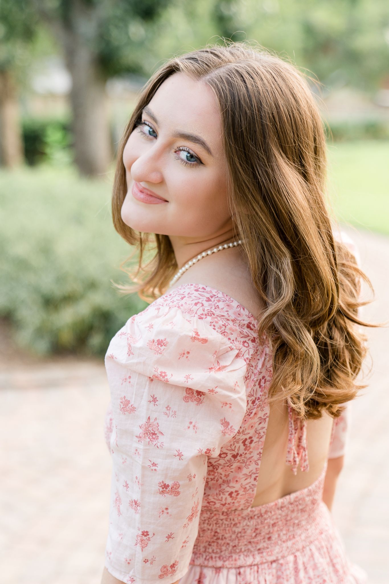 Young woman in pink floral off-shoulder dress with pearl necklace smiling outdoors in a garden setting.