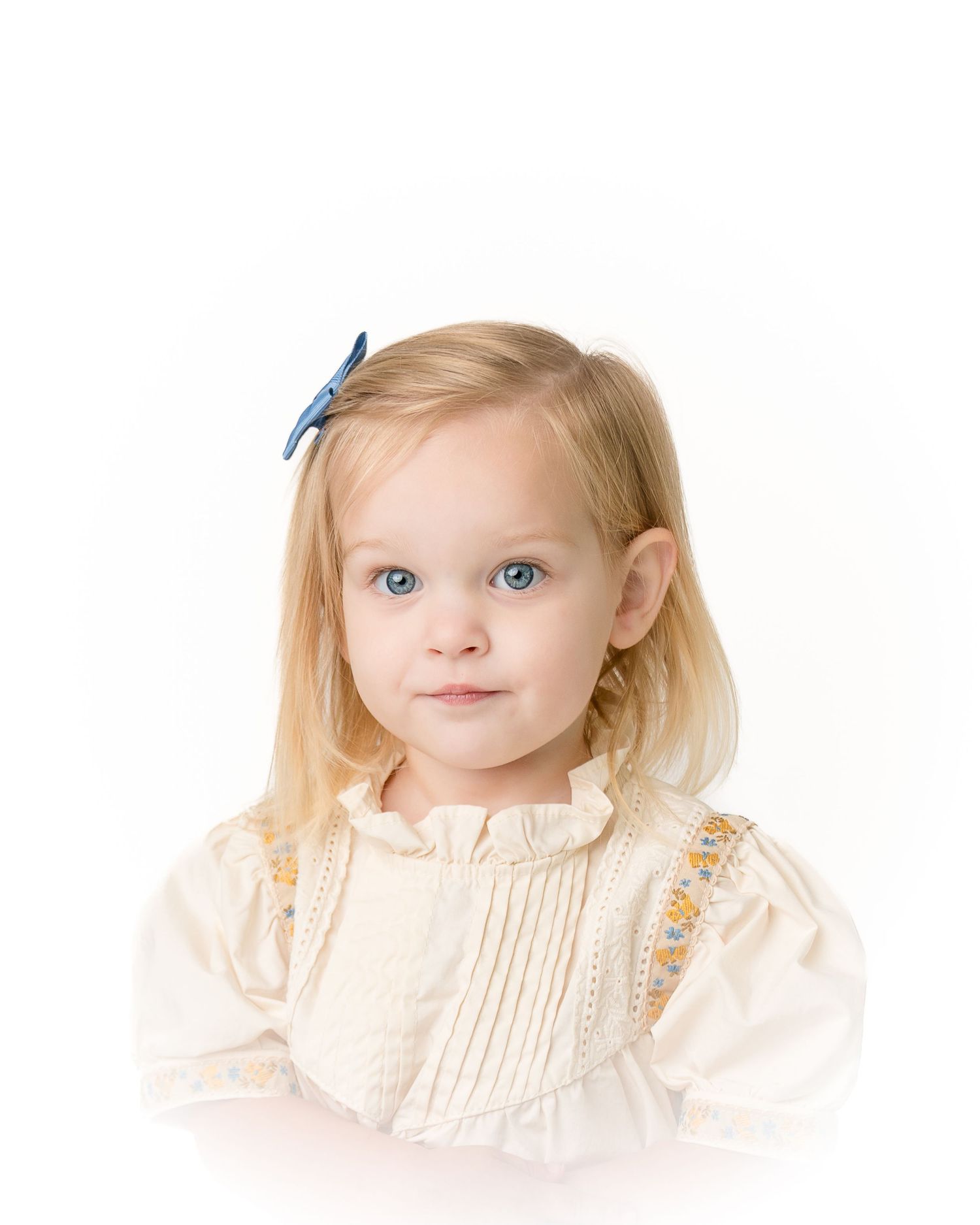 Close-up portrait of blonde toddler girl with blue eyes wearing cream smocked dress and blue hair bow.