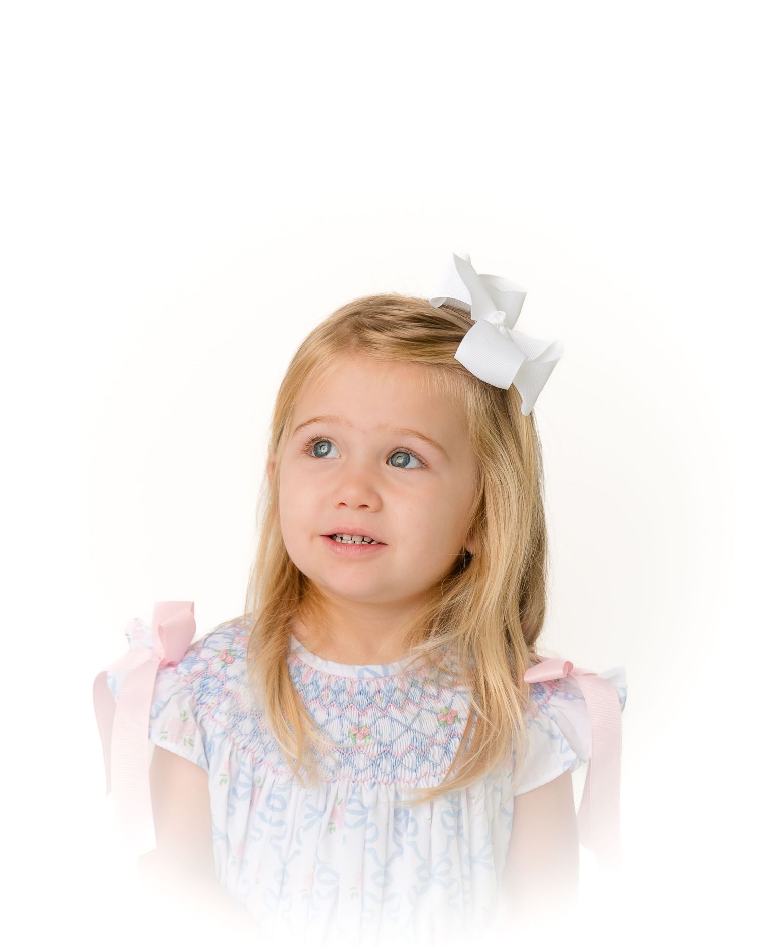 Blonde little girl with white hair bow in floral smocked dress looking upward.