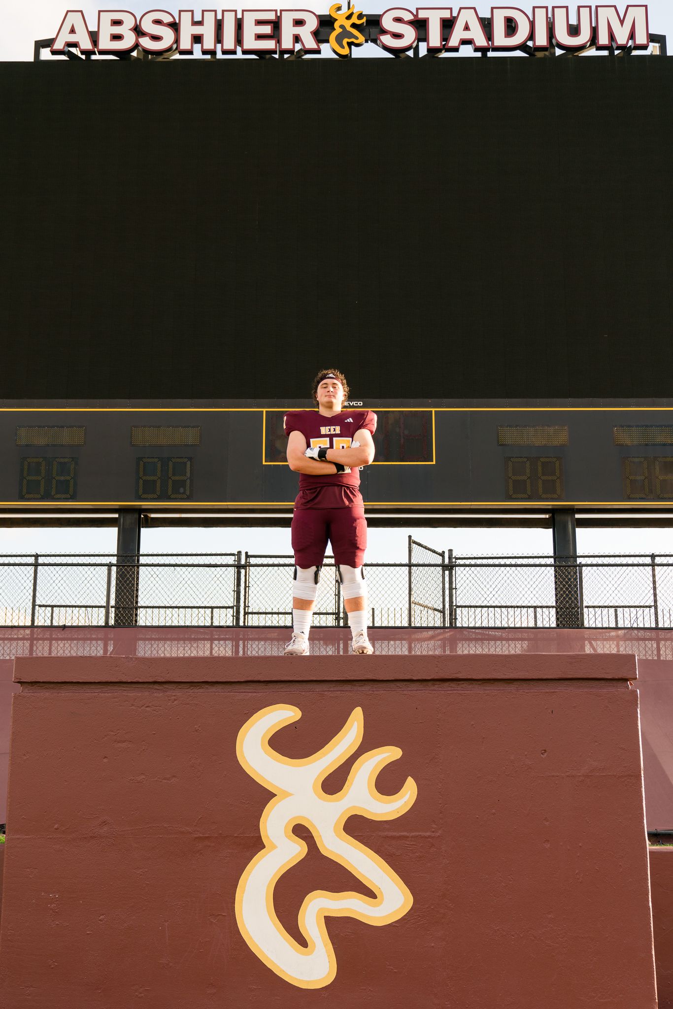 Football player in maroon uniform stands atop a wall at Abshier Stadium, posing confidently above a deer logo.