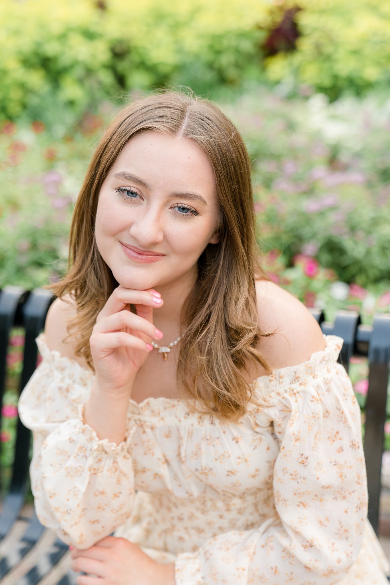 Young woman in floral off-shoulder dress poses gracefully in a garden, smiling softly with hand near chin.