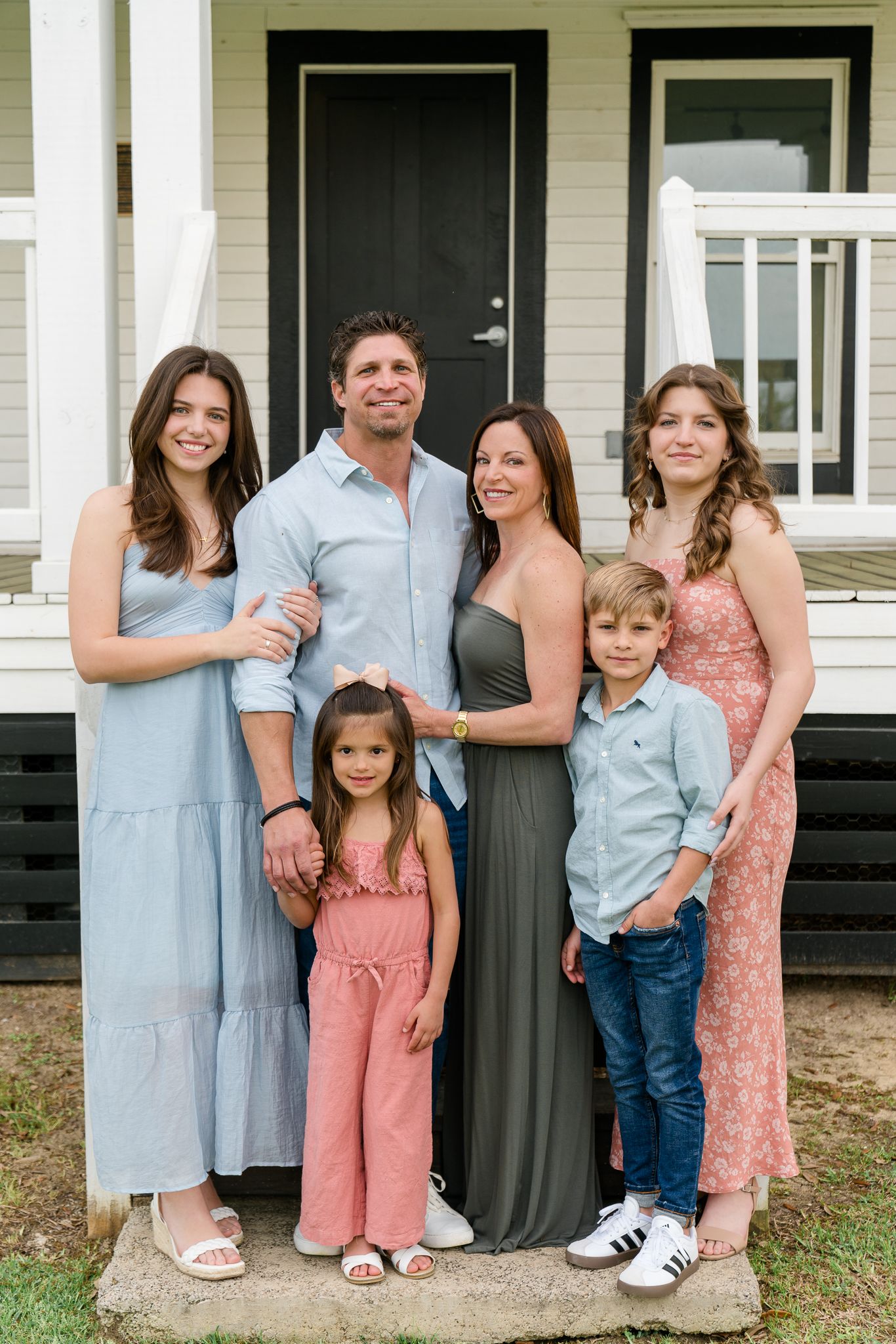 Family of seven smiling together on front porch steps wearing coordinated blue, gray and pink casual outfits.