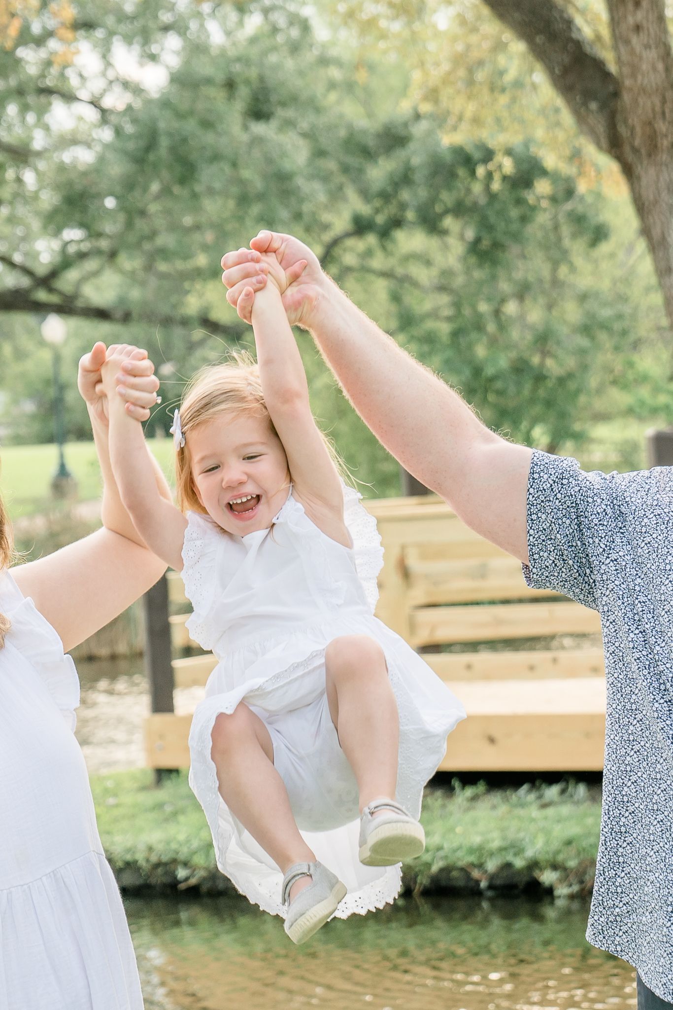 Two parents joyfully lift their laughing toddler girl in a white dress outdoors near a wooden dock.